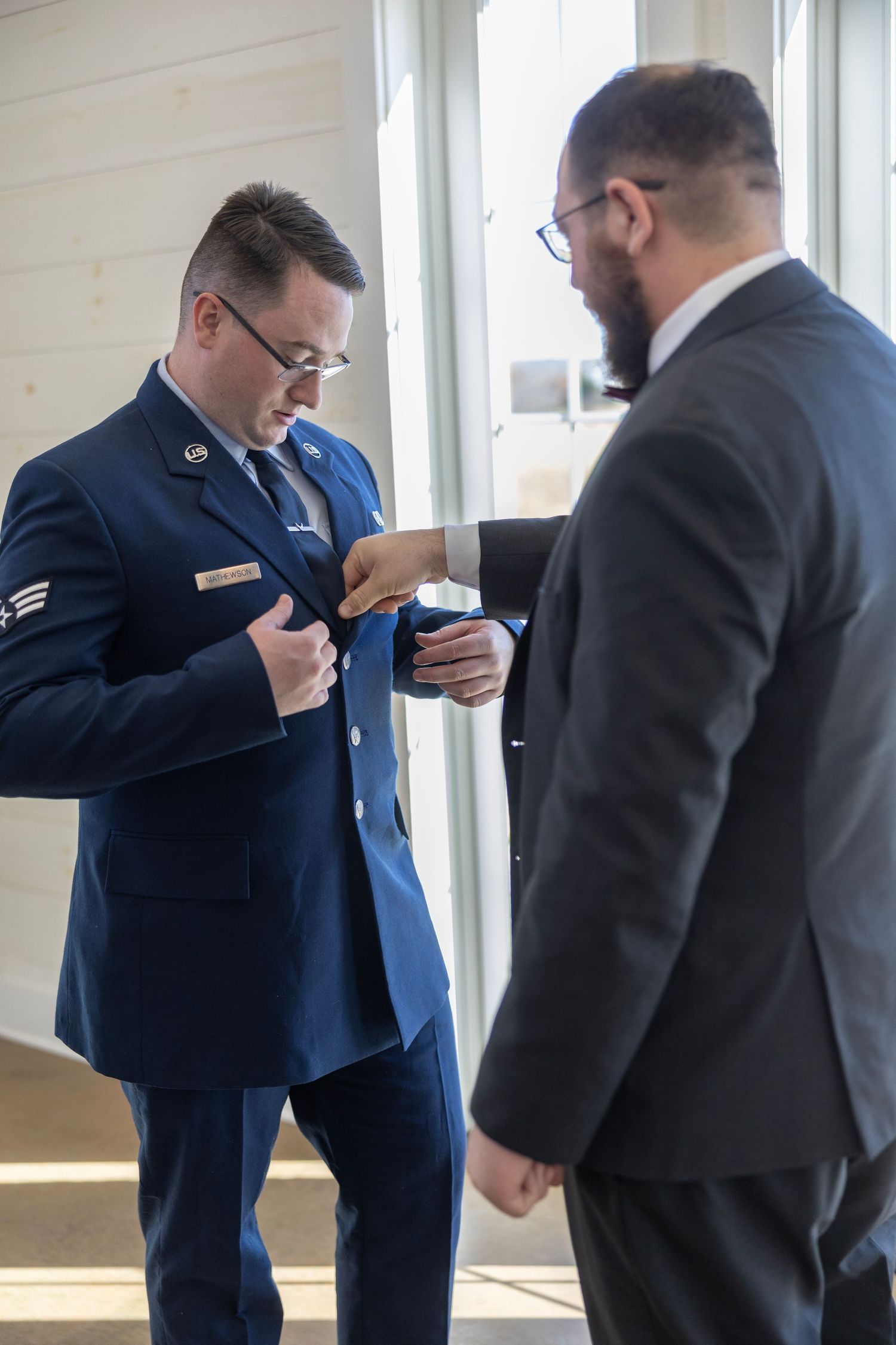 Two men in formal attire examine documents together by a window, one in military dress blues and one in a business suit.