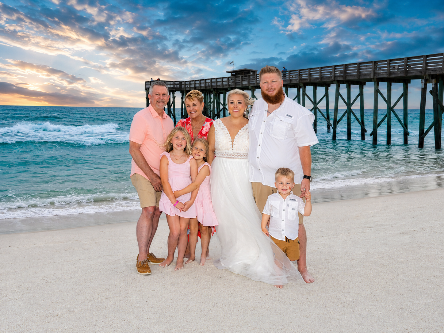 Family poses with newlyweds on a beach at sunset near a pier, children in pink dresses, groom in white shirt.