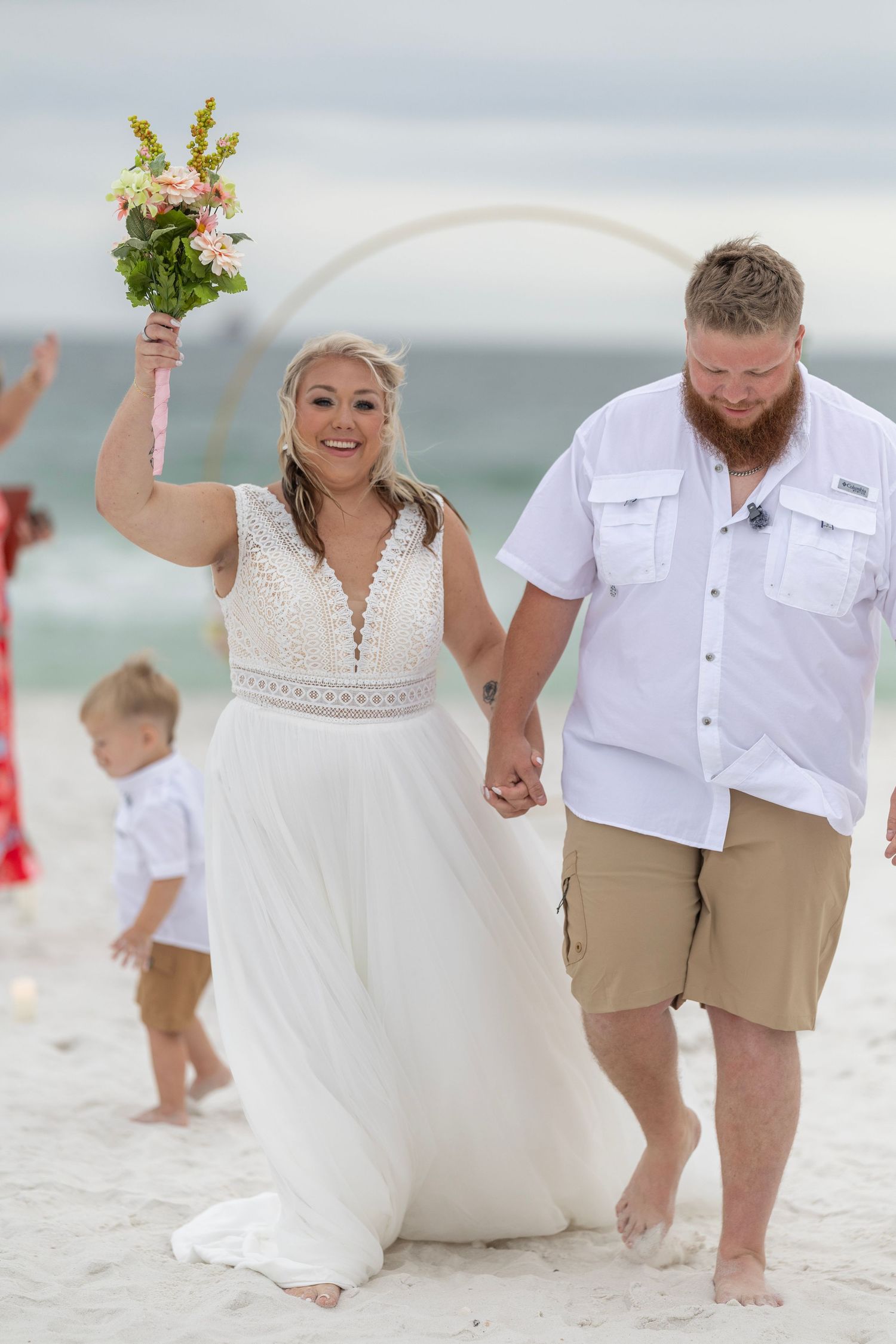 Joyful newlyweds holding hands on a sandy beach, bride in white gown raising bouquet, groom in white shirt and khaki shorts.