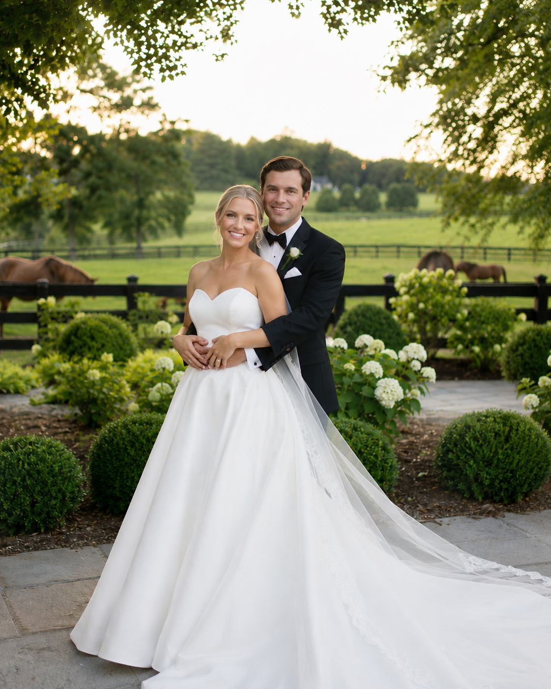 Bride in strapless white ball gown and groom in black tuxedo pose together at outdoor wedding venue with horses in background.