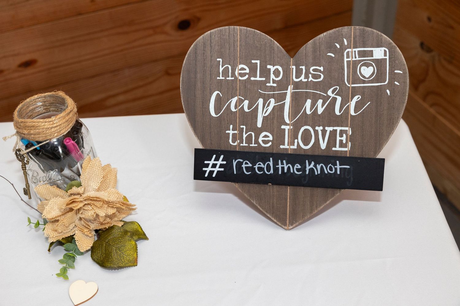 A rustic wooden heart-shaped wedding sign reads 'help us capture the love' with a camera icon and hashtag.