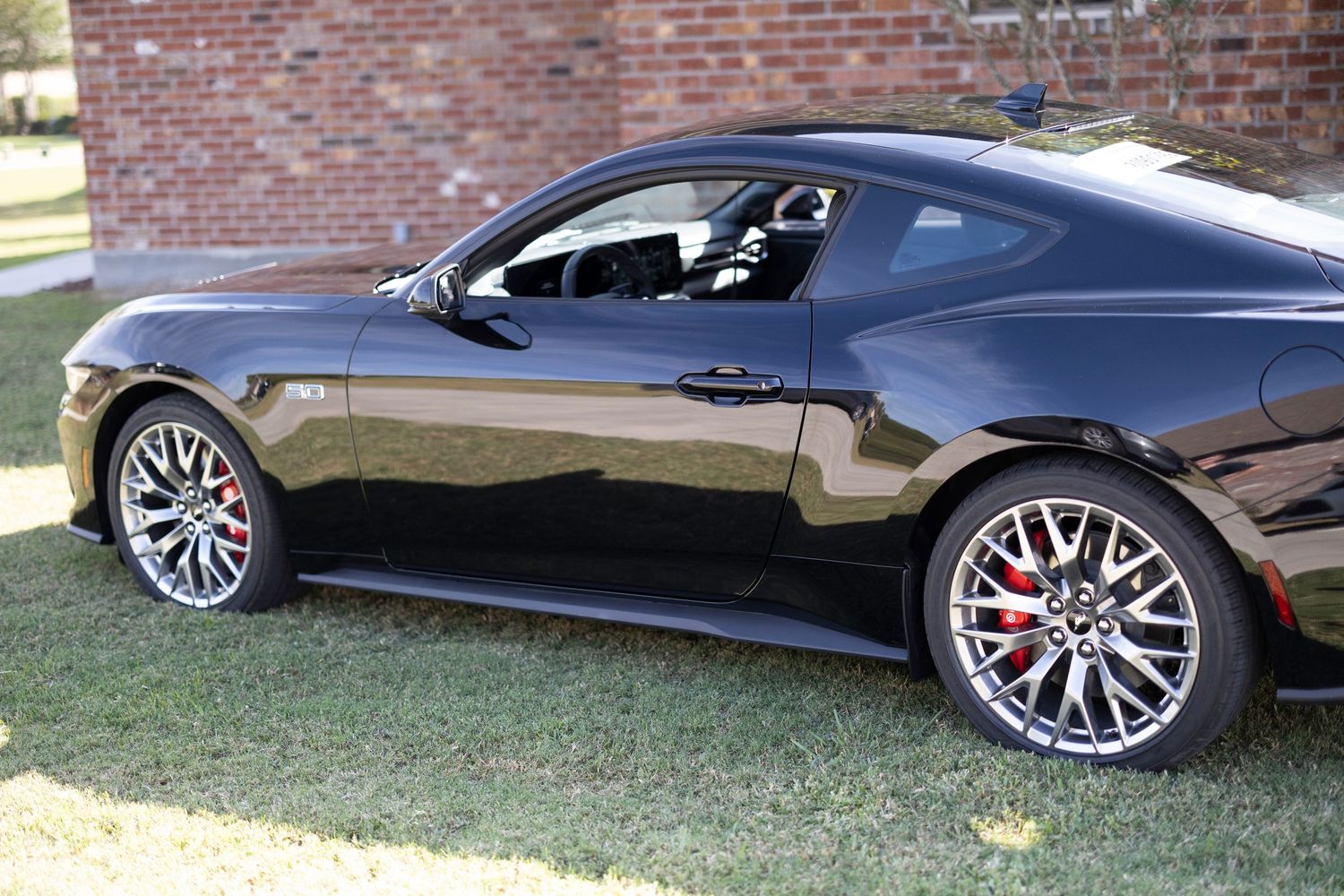 Side view of a sleek black sports car with custom wheels parked on grass near a brick wall.
