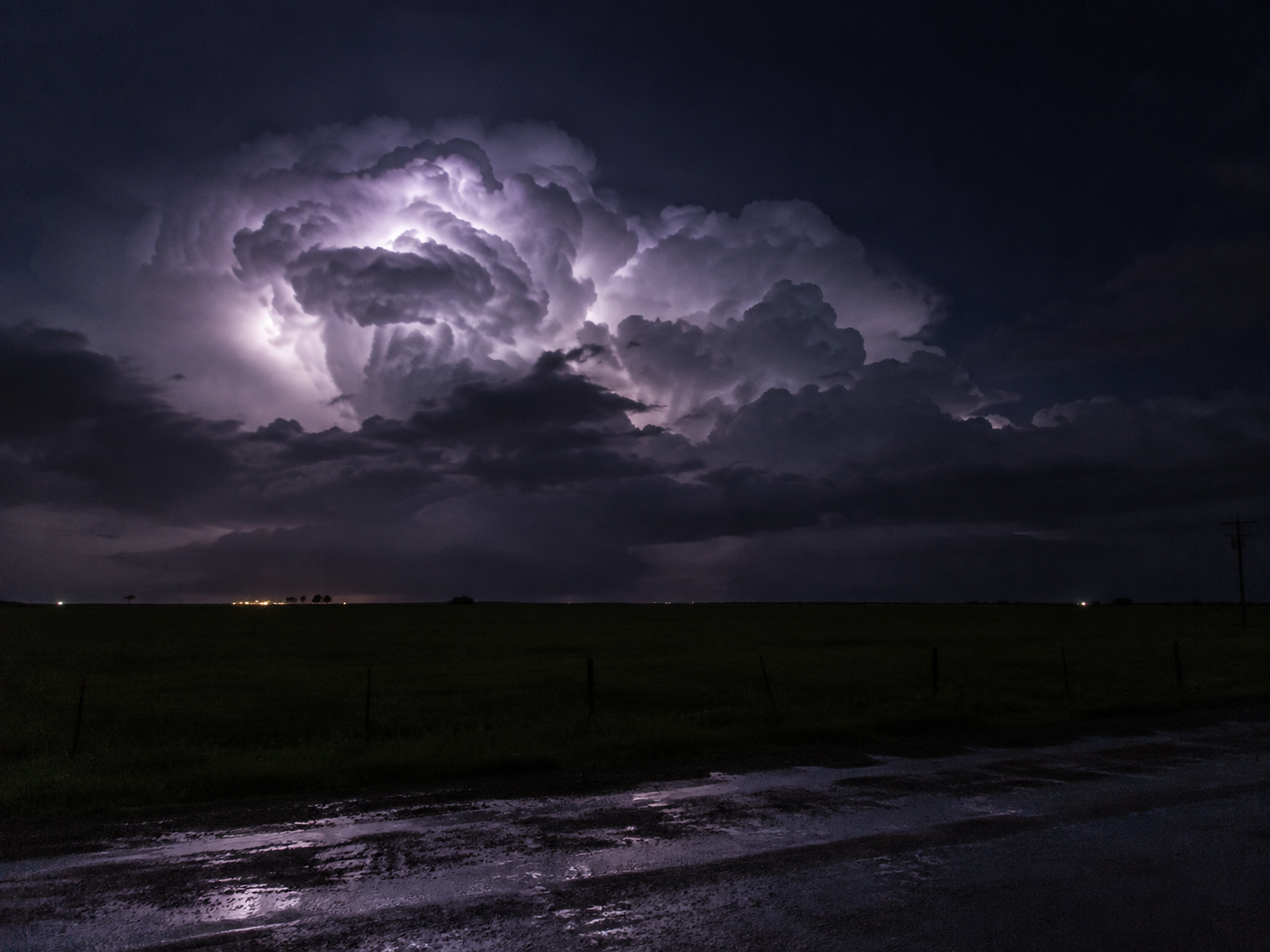 Lightning illuminates massive storm clouds over a flat rural landscape at night, reflecting on a wet road.