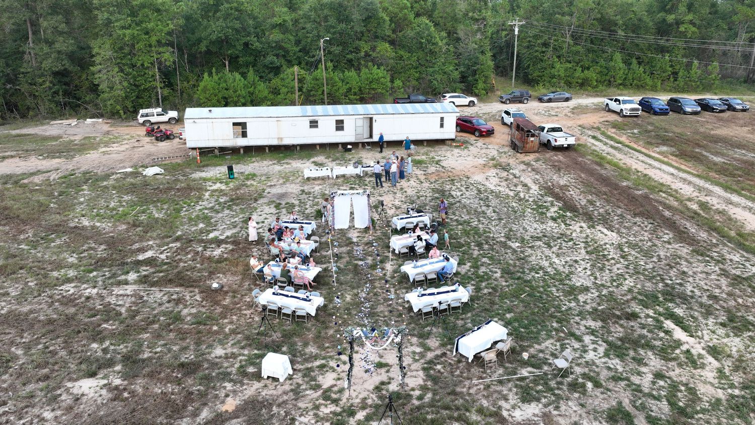 Aerial view of an outdoor gathering with white tables and chairs near a mobile office building surrounded by pine trees.