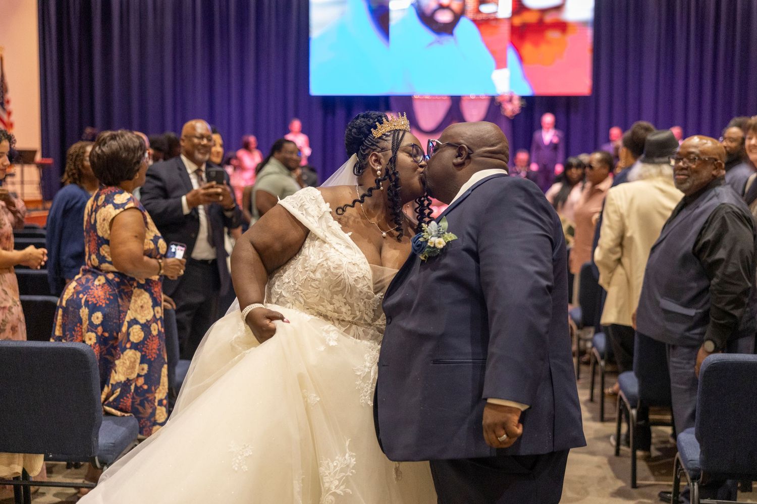 Bride and groom share a kiss at their wedding ceremony in a decorated venue with guests celebrating around them.