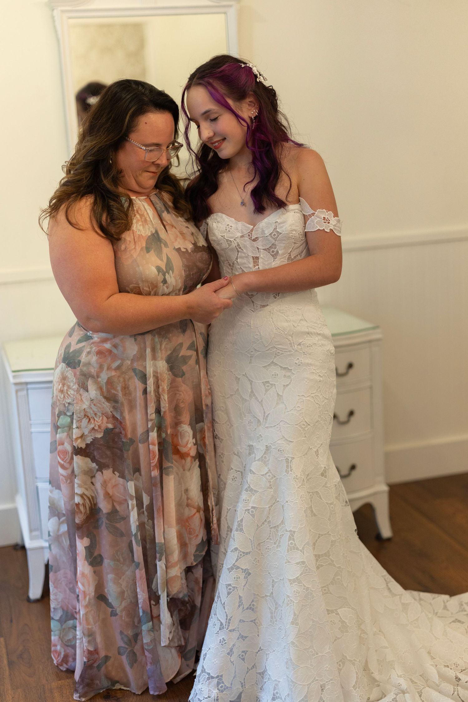 A bride in a white lace gown with purple hair shares a tender moment with a woman in a floral dress.