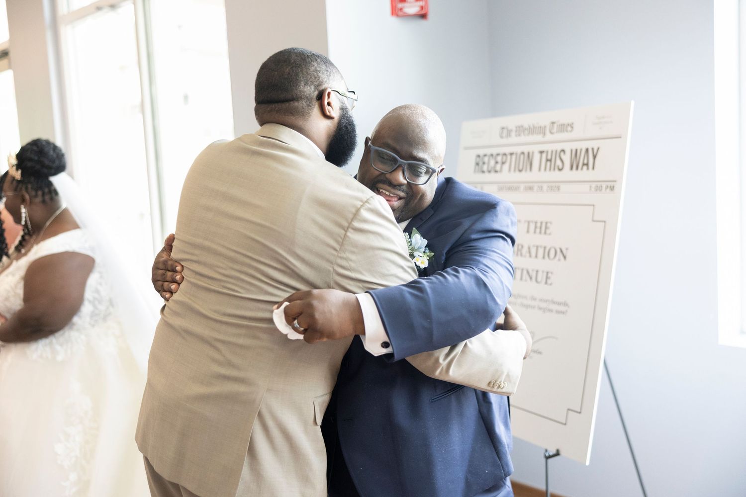 Two men share a warm hug at a wedding reception, with a bride visible in the background near a directional sign.