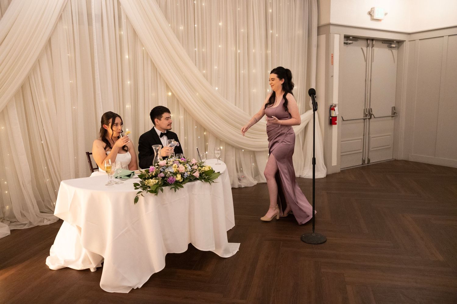 Woman giving a wedding toast at a microphone while bride and groom sit at sweetheart table with floral centerpiece.