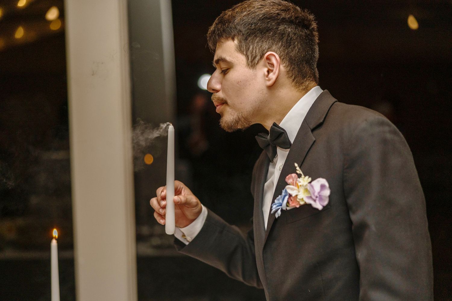 Groom in gray suit with floral boutonniere blowing out a candle at a wedding reception.