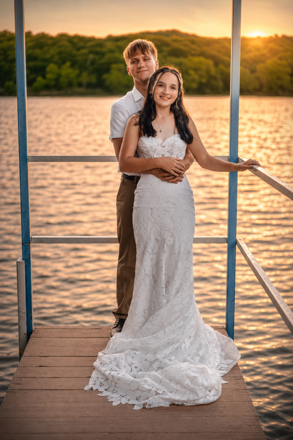 Bride in white lace gown and groom embrace on a lakeside dock at golden sunset.