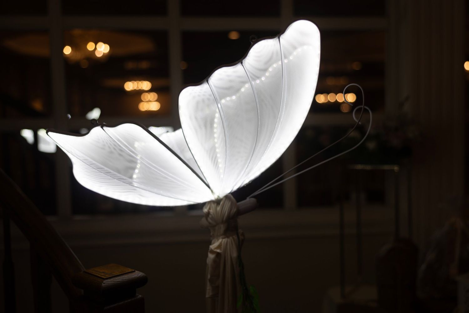Glowing white butterfly sculpture lamp illuminated in a dark room with warm bokeh lights in the background.