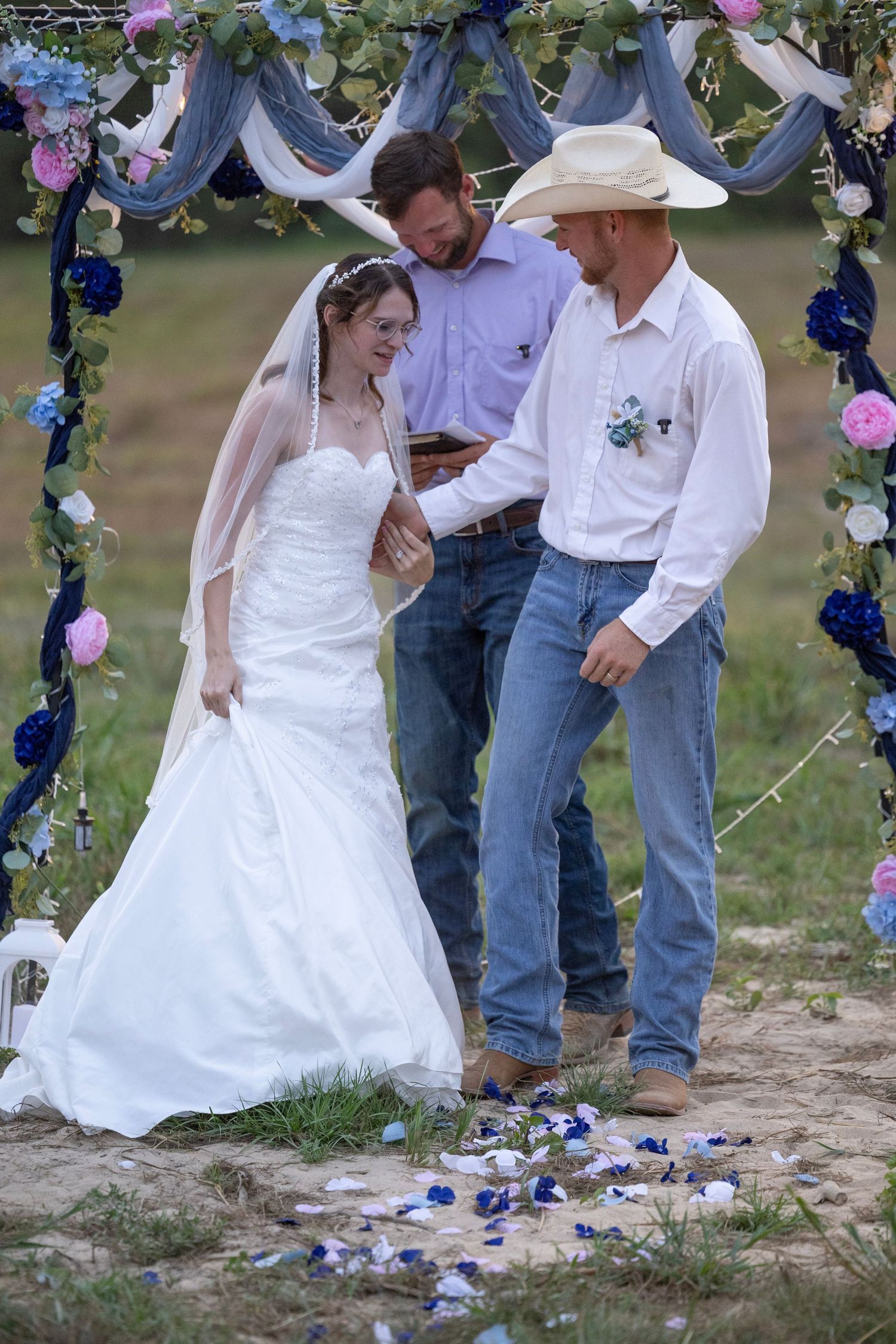 Outdoor country wedding ceremony with bride in white gown and groom in cowboy hat under floral arch.