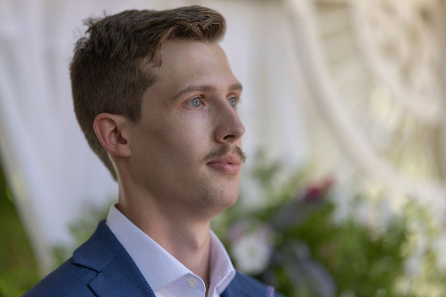 Young man with mustache in blue suit gazing thoughtfully at outdoor wedding ceremony with floral backdrop.