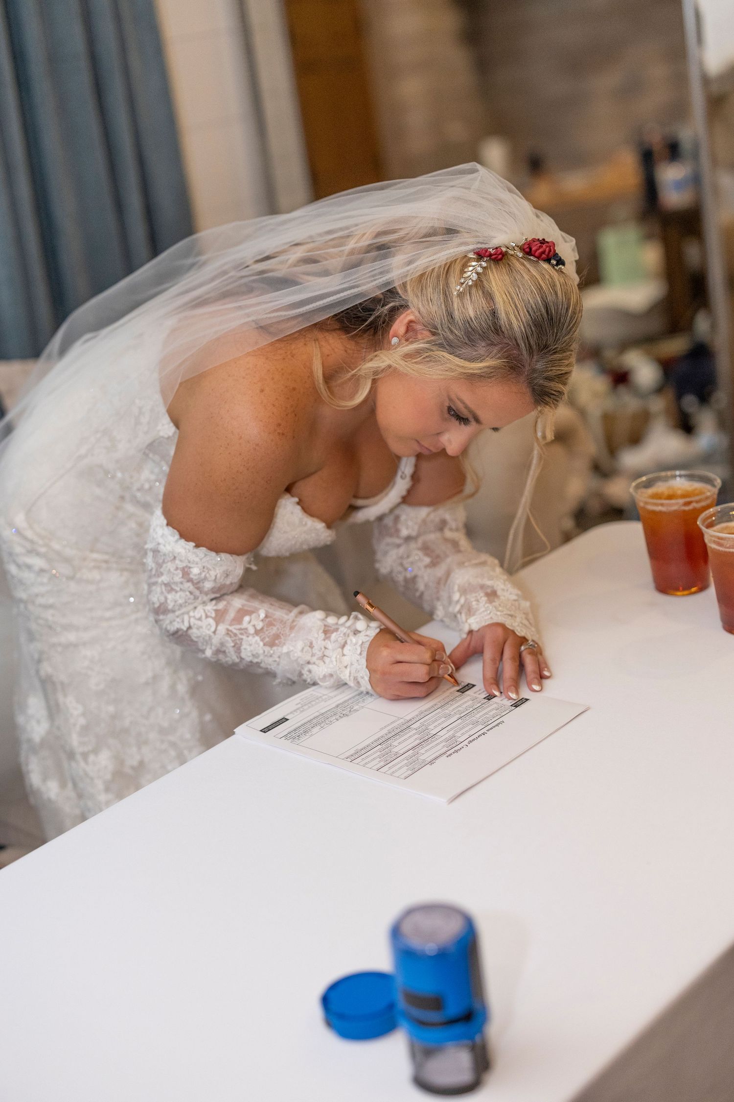 A bride in white lace dress signs marriage documents at a table while leaning over the paperwork.