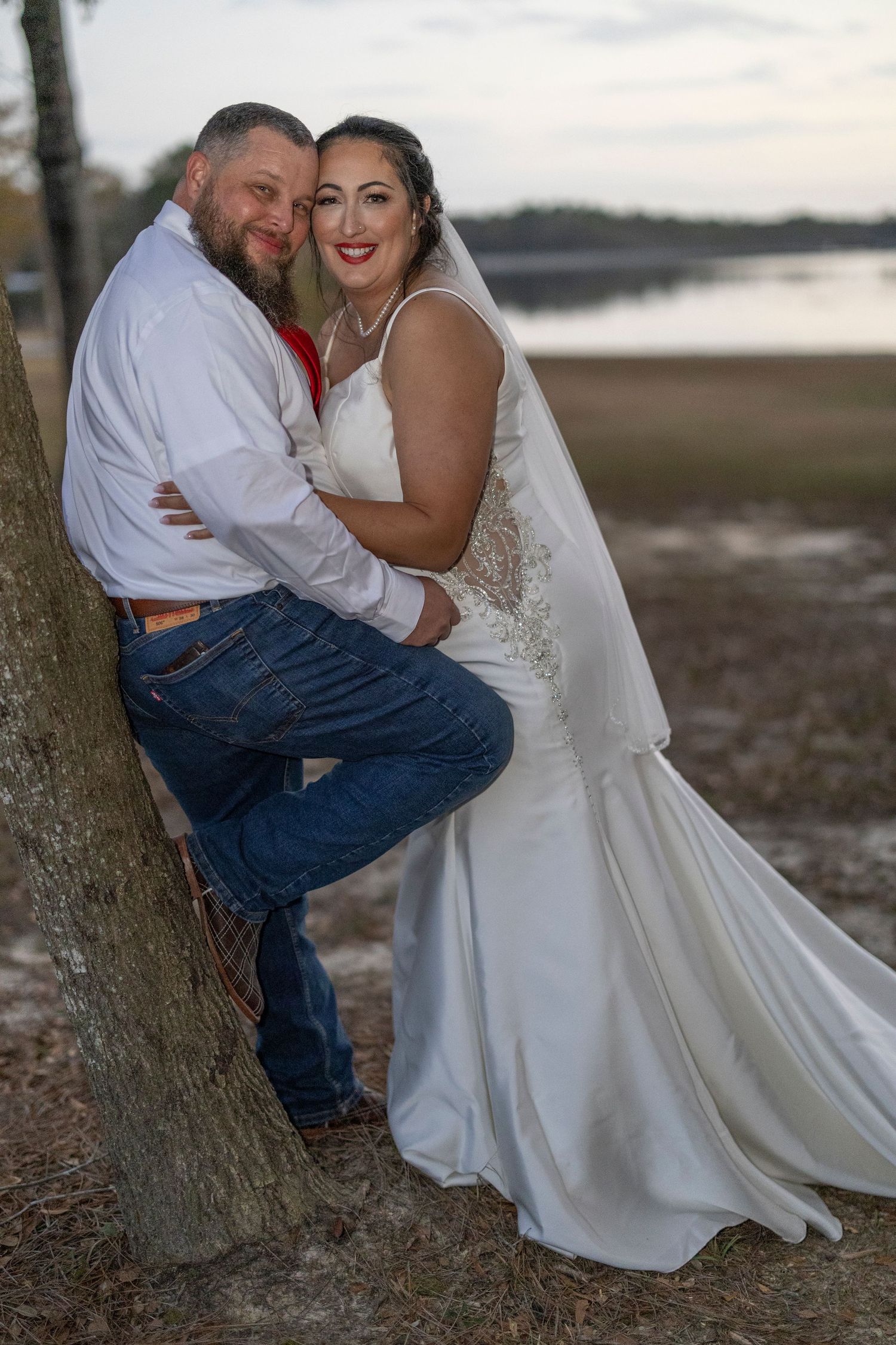 A bride and groom share intimate moments during their outdoor wedding photoshoot by a tree at sunset.