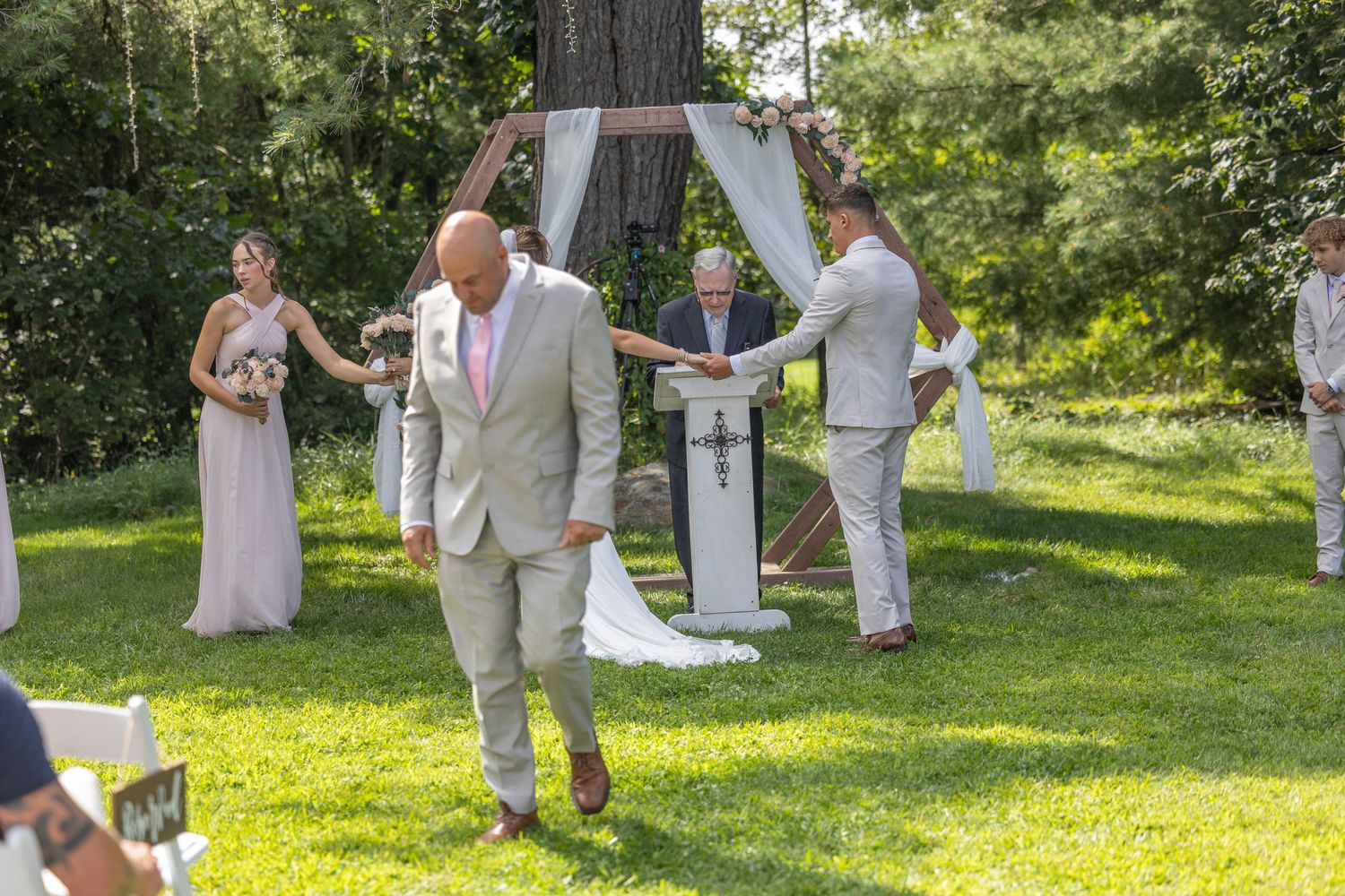 Wedding party gathers under tall trees in a sunlit garden setting for outdoor celebration photos.