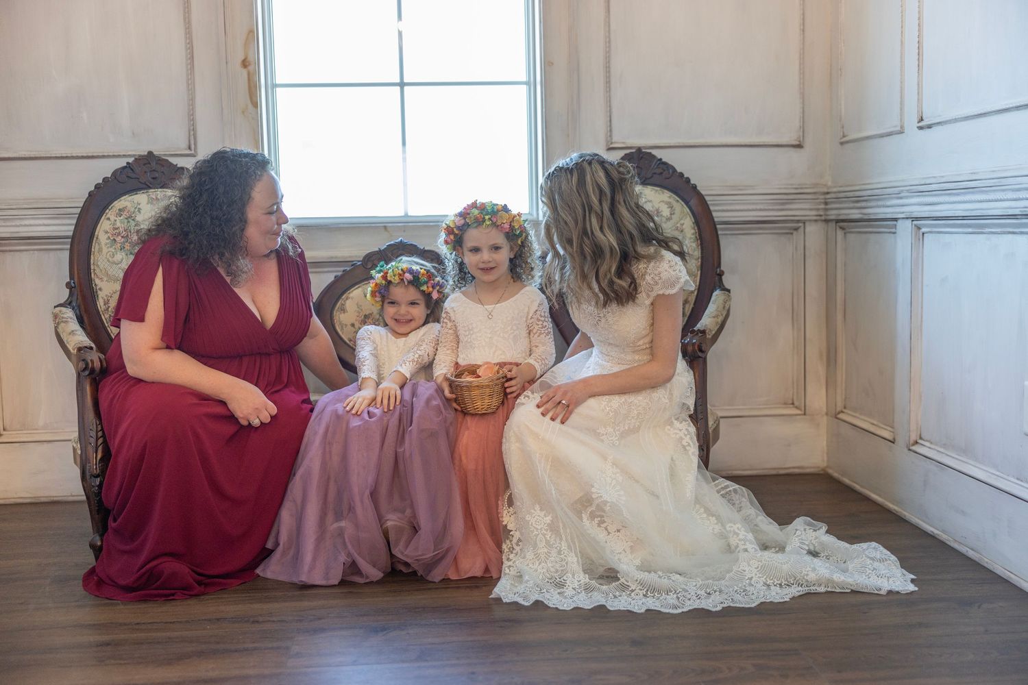 A group sits together on an elegant vintage sofa wearing formal dresses in burgundy, pink and white.