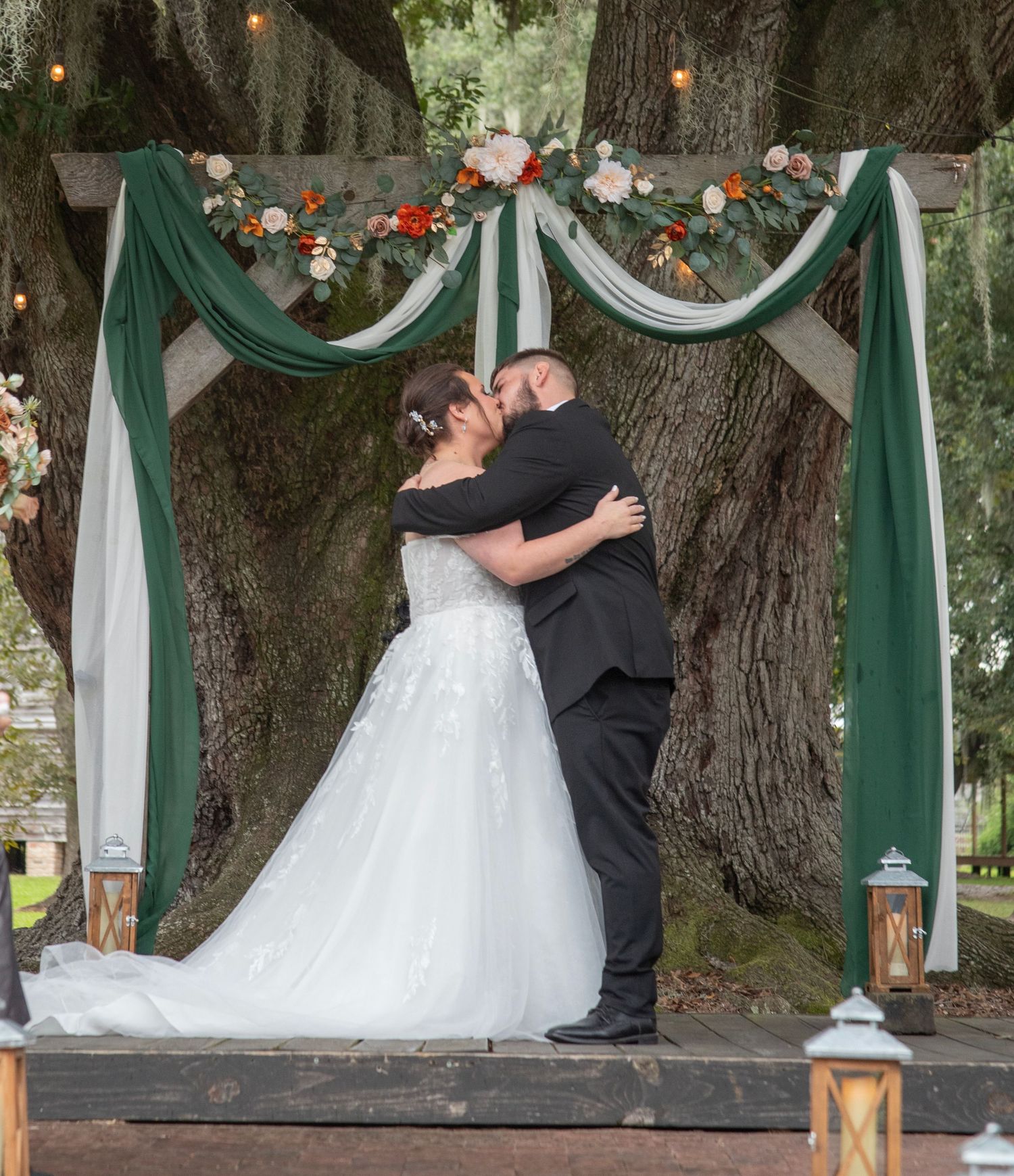 A romantic wedding ceremony moment captured under trees decorated with green and white fabric draping and lanterns.