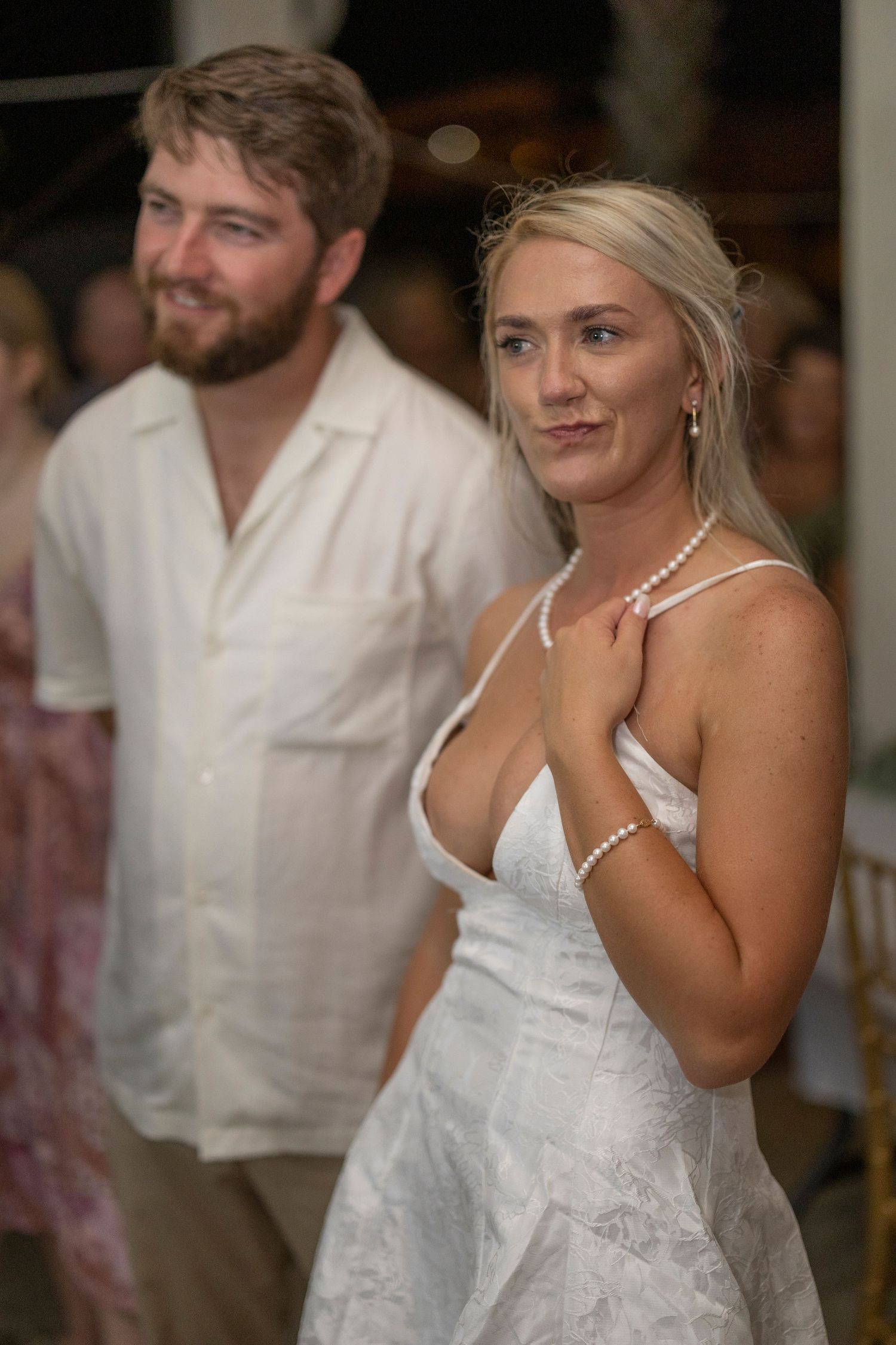 A bride in a white lace dress and pearl necklace stands next to someone wearing a white shirt at a wedding reception.