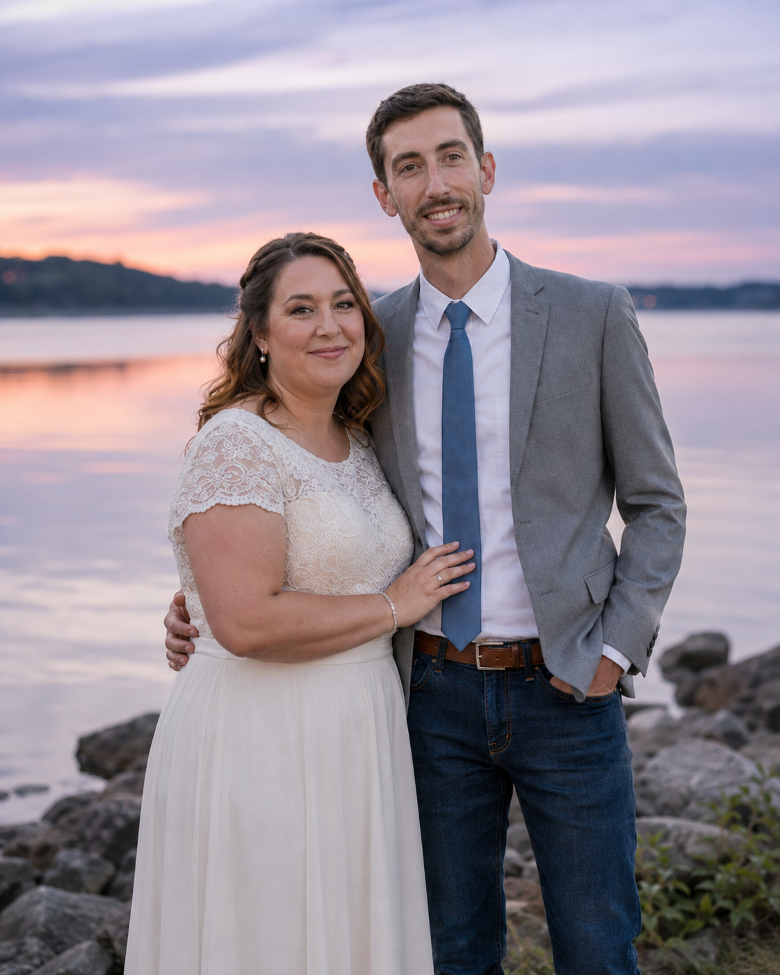 Wedding couple embraces by rocky waterfront at sunset with colorful pink and purple sky reflecting on water.