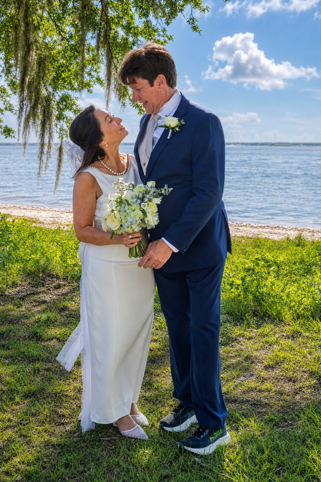 Bride and groom share a romantic moment by the waterfront, she in white gown holding bouquet, he in navy suit.