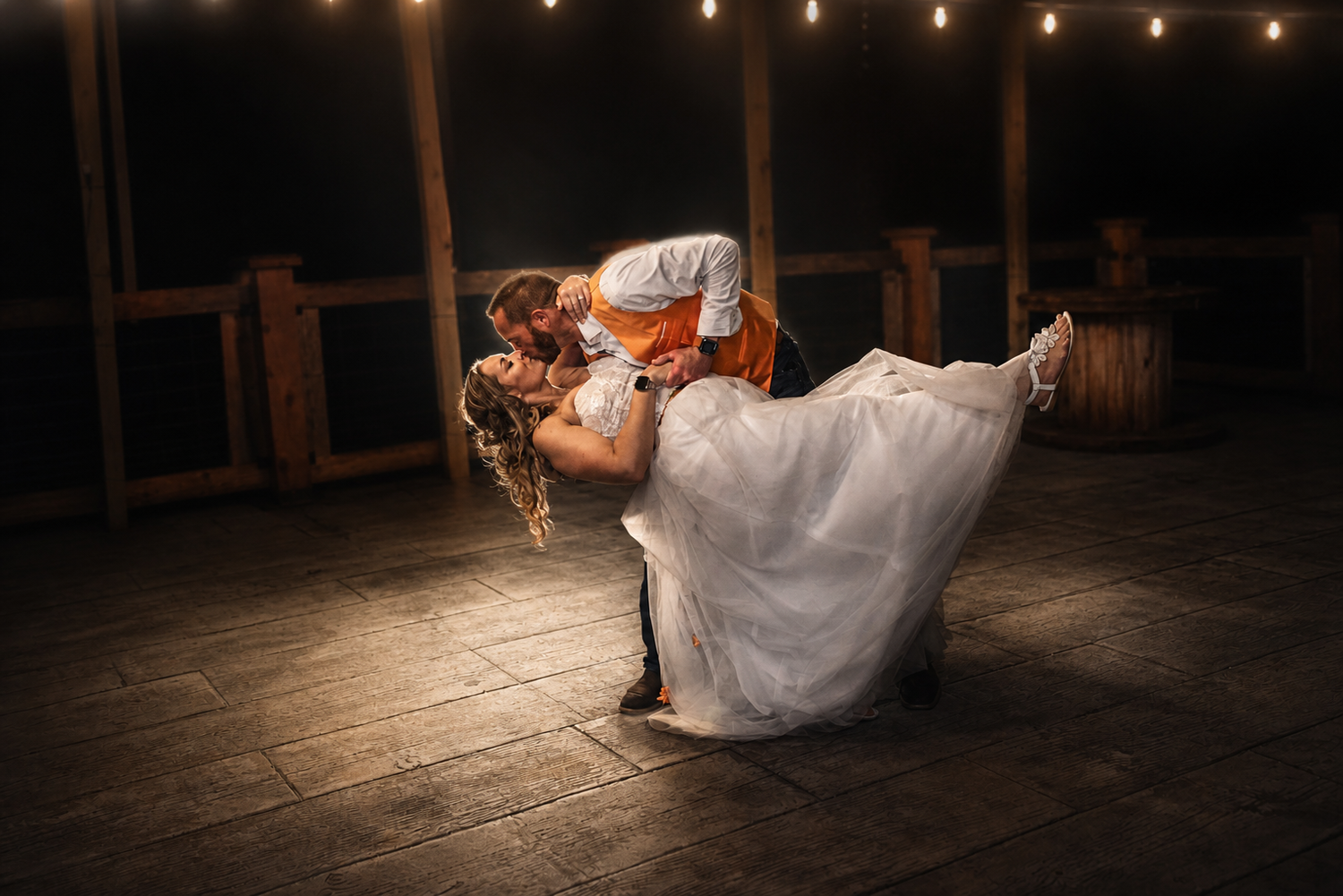 Bride and groom share romantic first dance dip kiss under warm string lights on rustic wooden dance floor.