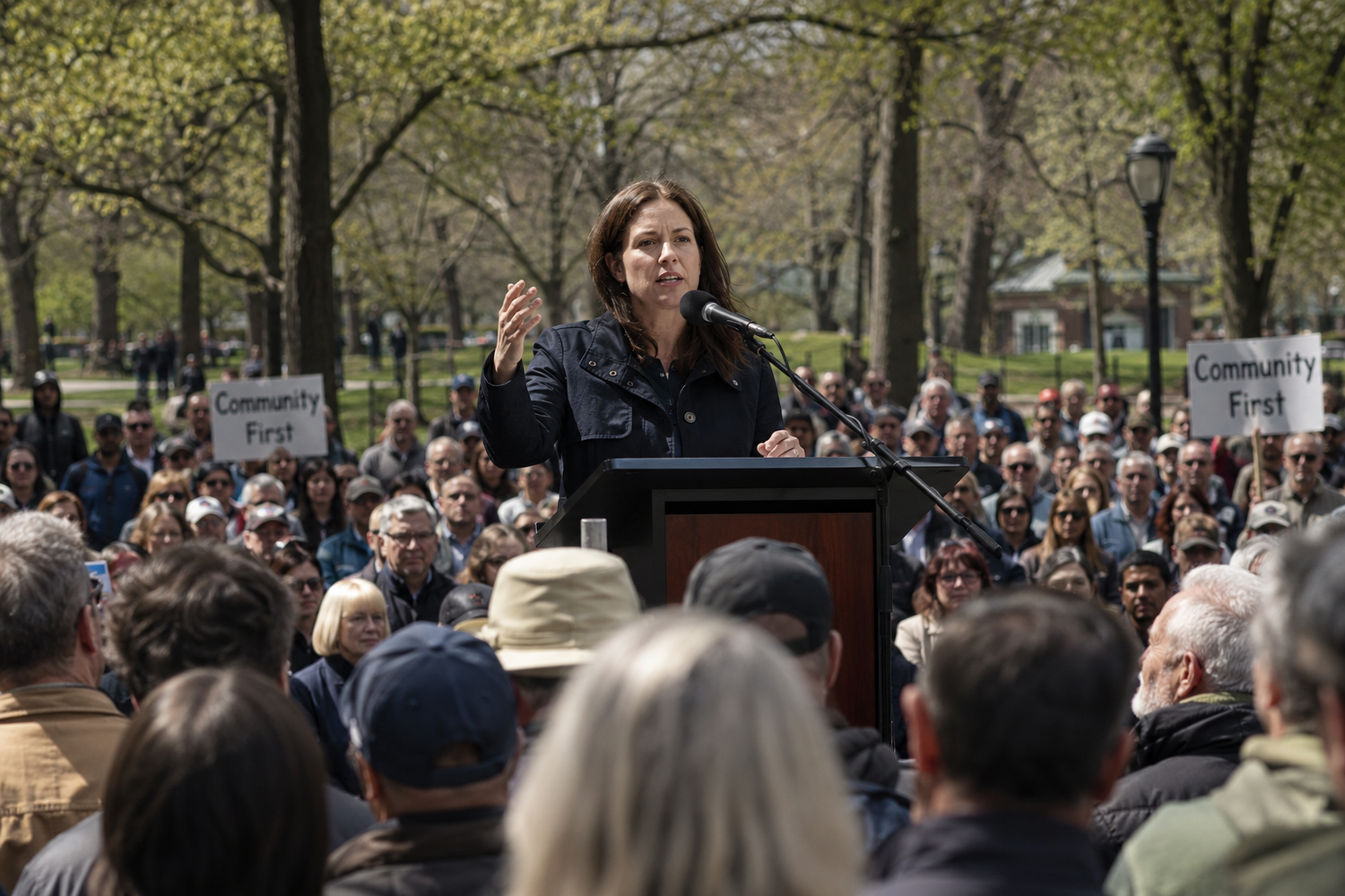 Woman speaks at outdoor podium to large crowd holding Community First signs in a park rally.