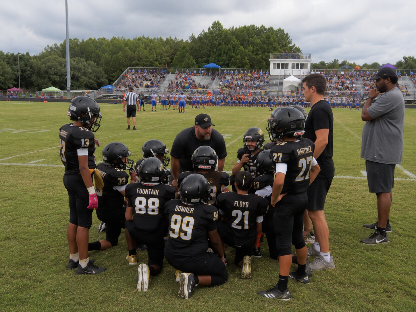 Youth football team huddles with coaches on green field, stadium crowd visible in background during game day.