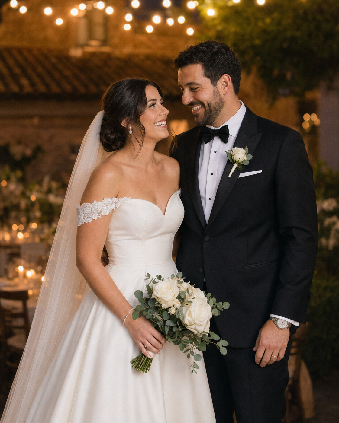Smiling bride in off-shoulder gown and groom in black tux share a glance at candlelit outdoor wedding reception.