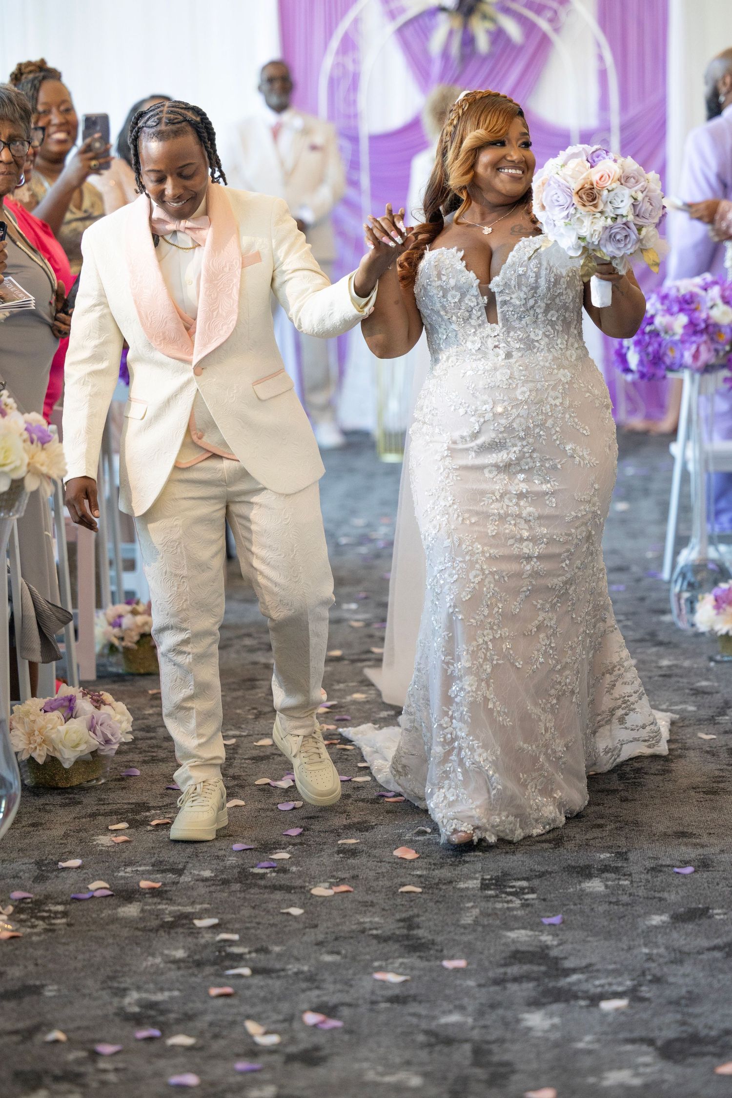 Newlywed couple walks down the aisle, bride in lace gown holding bouquet, groom in cream tuxedo, guests celebrating.