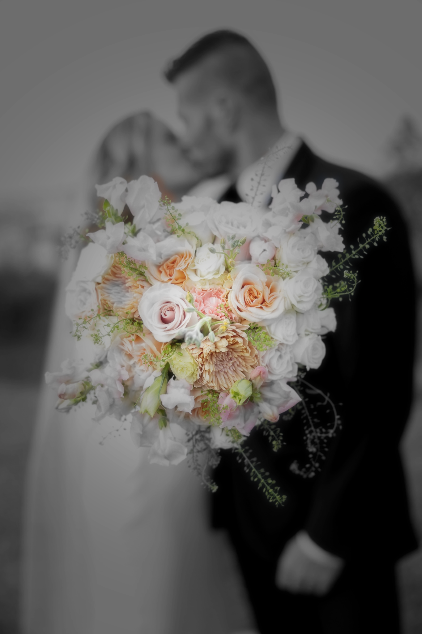Close-up of a colorful bridal bouquet with roses with blurred bride and groom kissing in black and white.