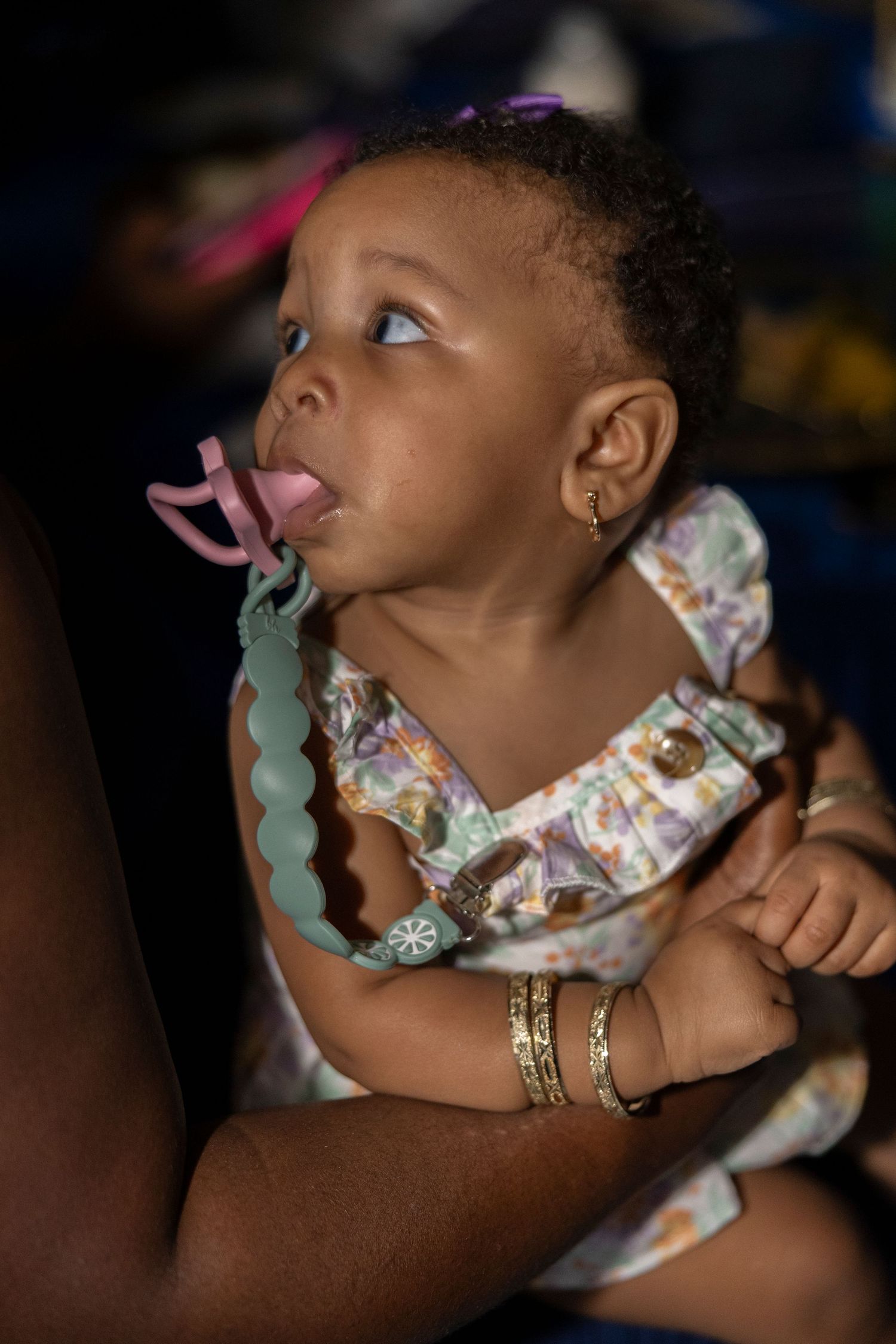 Adorable baby girl with pacifier and earrings, wearing floral dress, held by adult with gold bracelets.