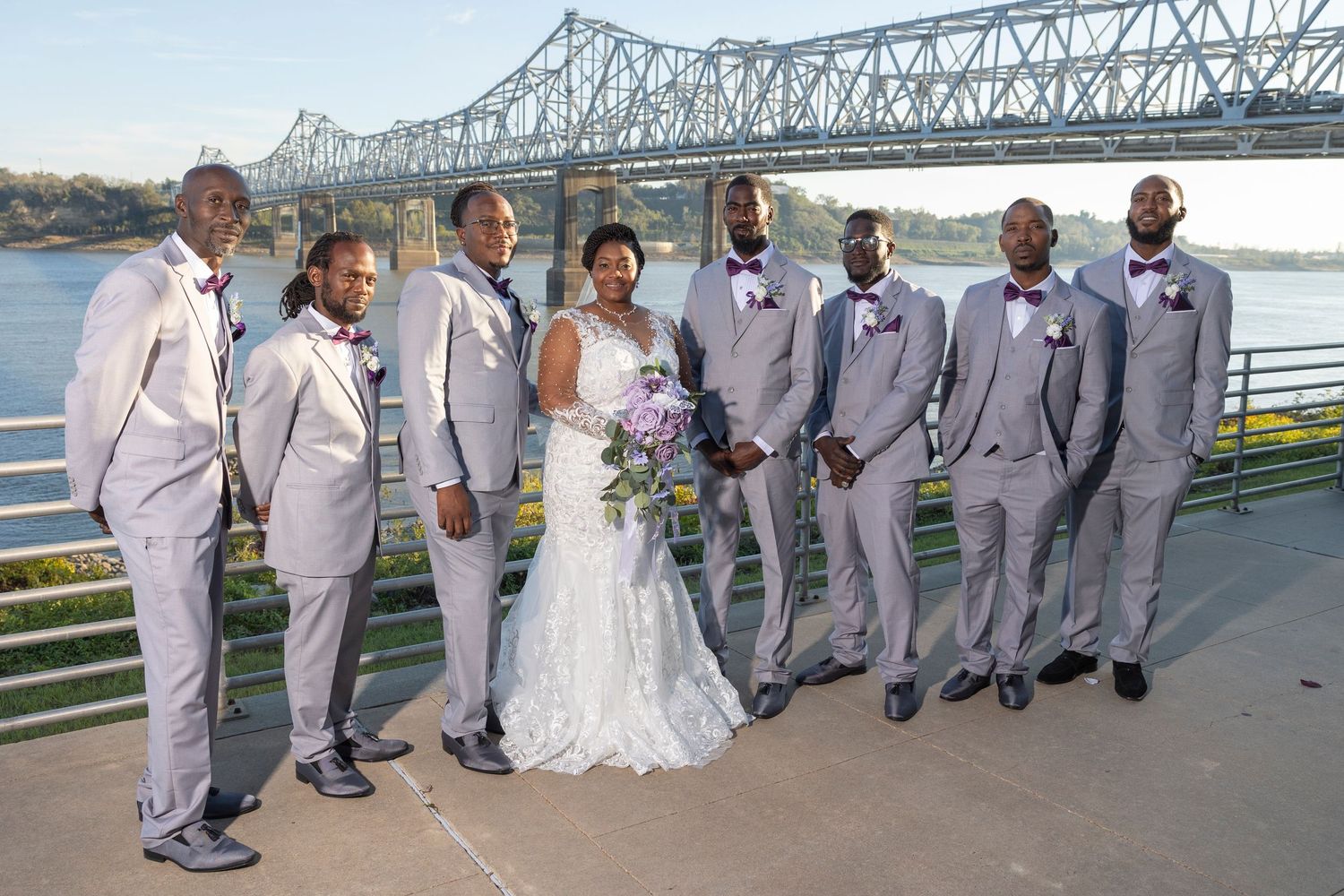 Bride in white lace gown poses with groomsmen in gray suits and burgundy bow ties near a river bridge.