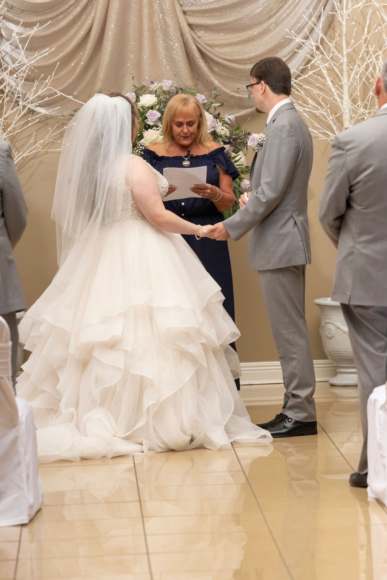 Wedding ceremony taking place with officiant and couple standing under floral arch with draped fabric backdrop.