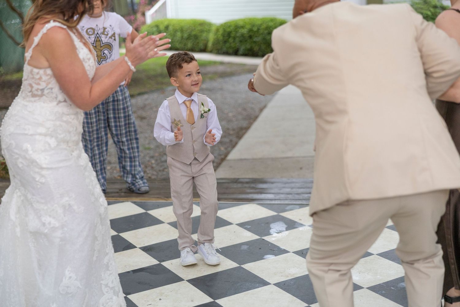 A young attendant in a light gray suit stands on a black and white checkered dance floor at a wedding celebration.