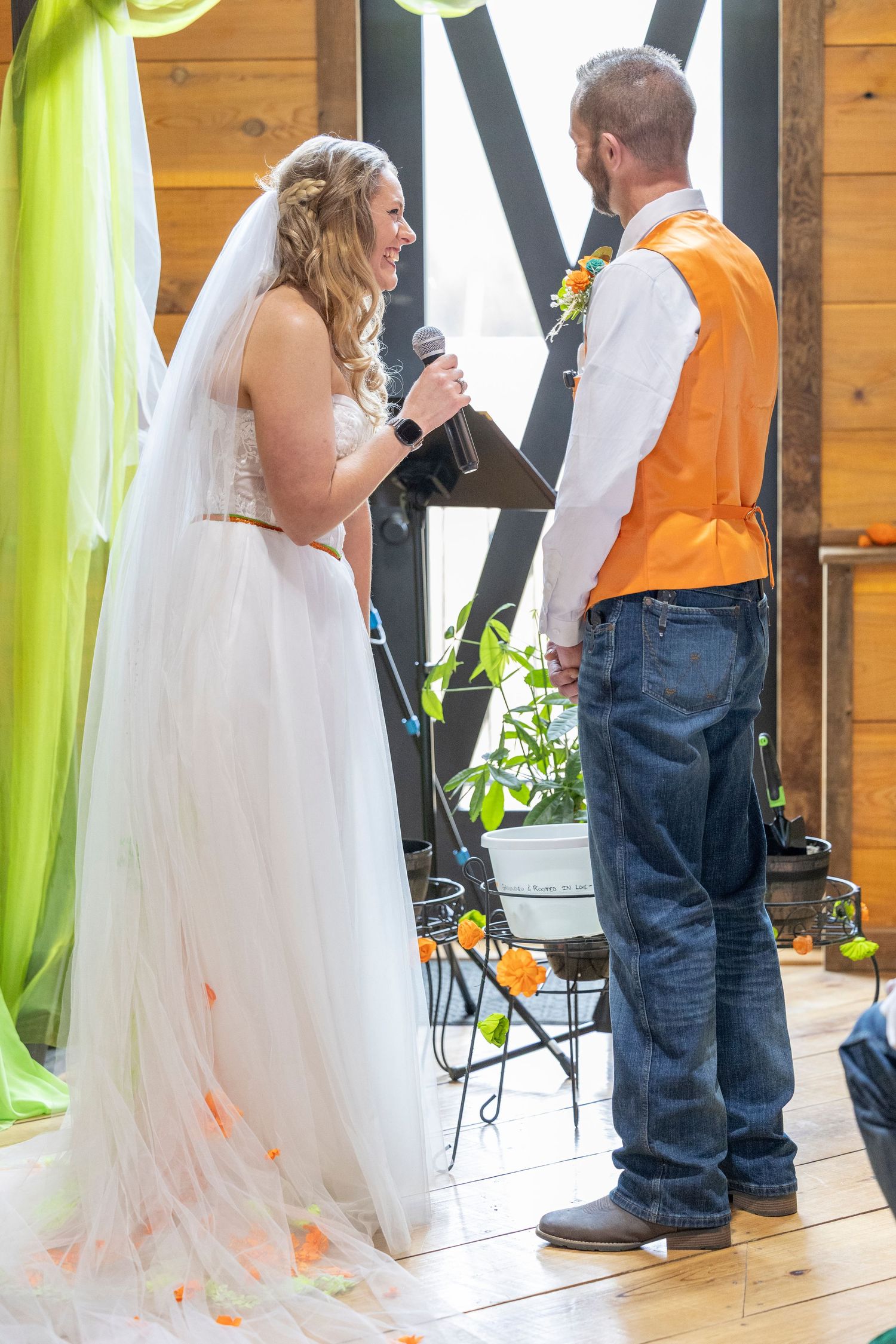 Bride in white gown reads vows to groom in orange vest and jeans during rustic barn wedding ceremony.