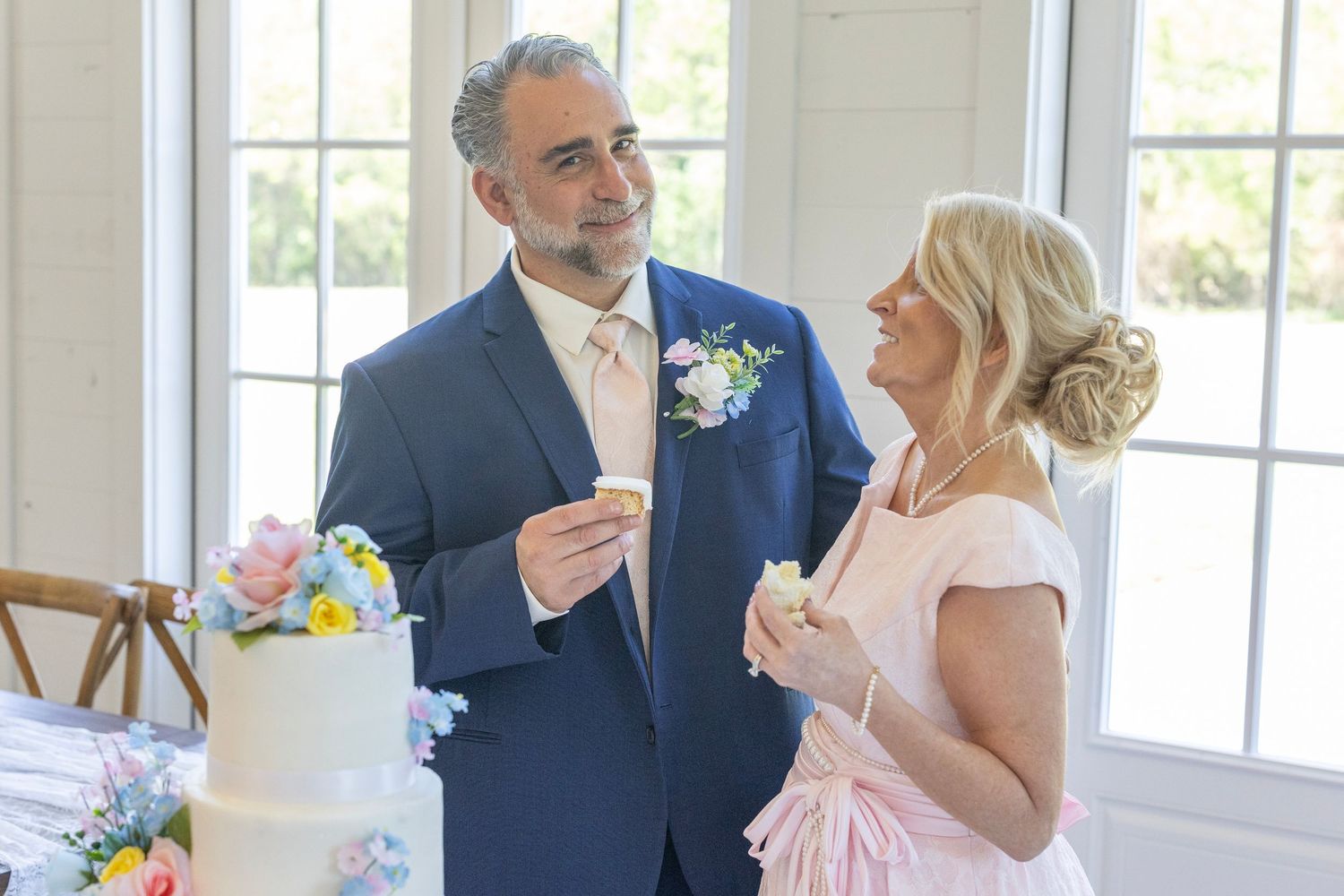 Couple sharing wedding cake at elegant reception, groom in navy suit with boutonniere, bride in pink dress.