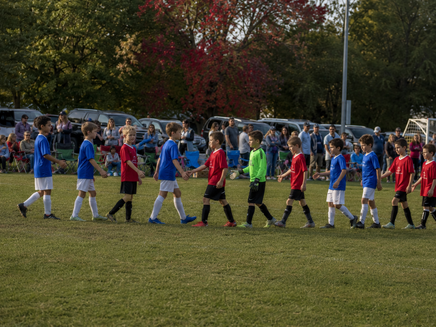 Youth soccer teams in red and blue uniforms shake hands on the field after an autumn game.