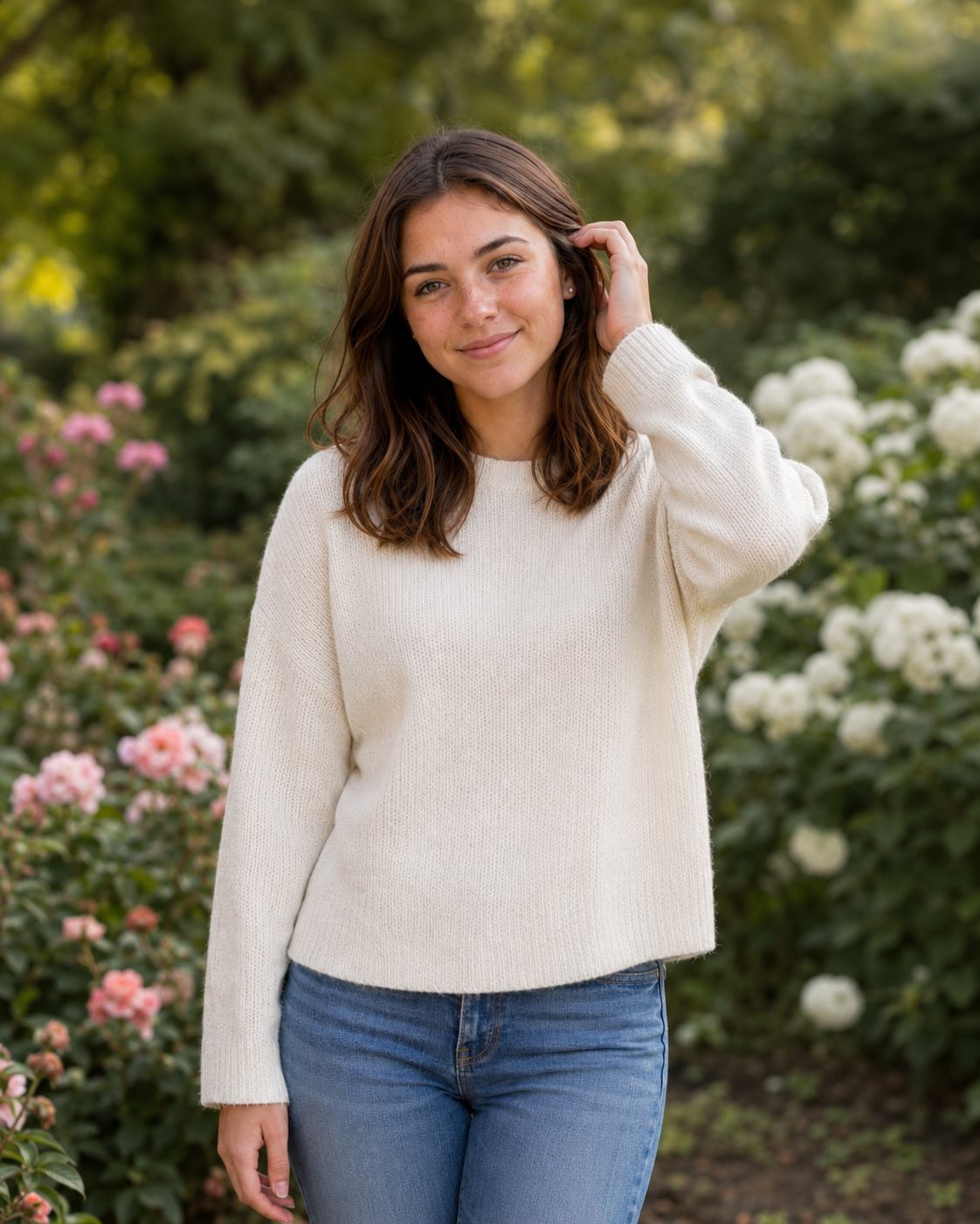 Smiling woman in ivory sweater posing among blooming rose bushes in garden setting.