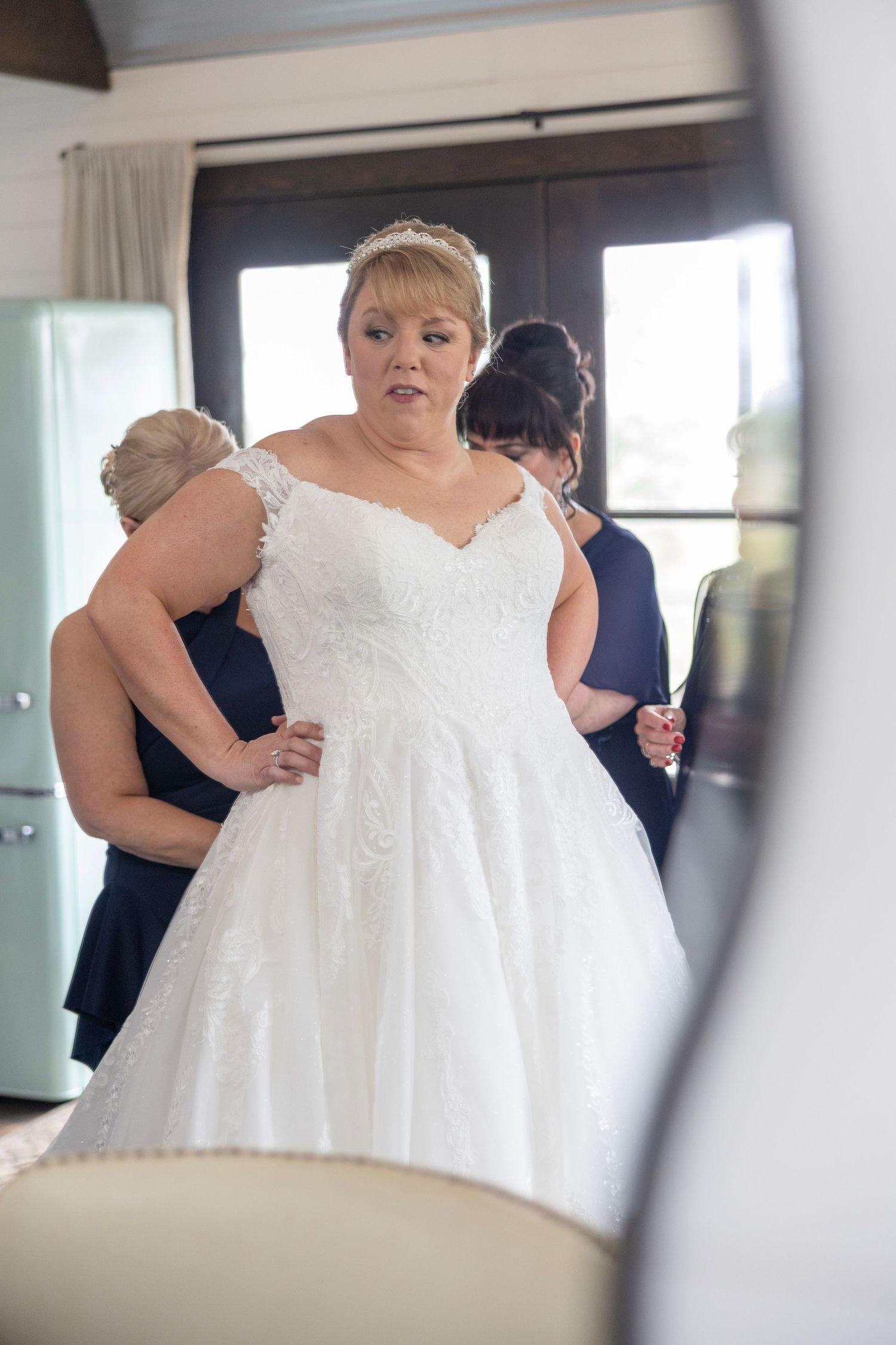Bride in white lace ball gown wedding dress being helped by bridesmaids in navy blue dresses during bridal preparations.