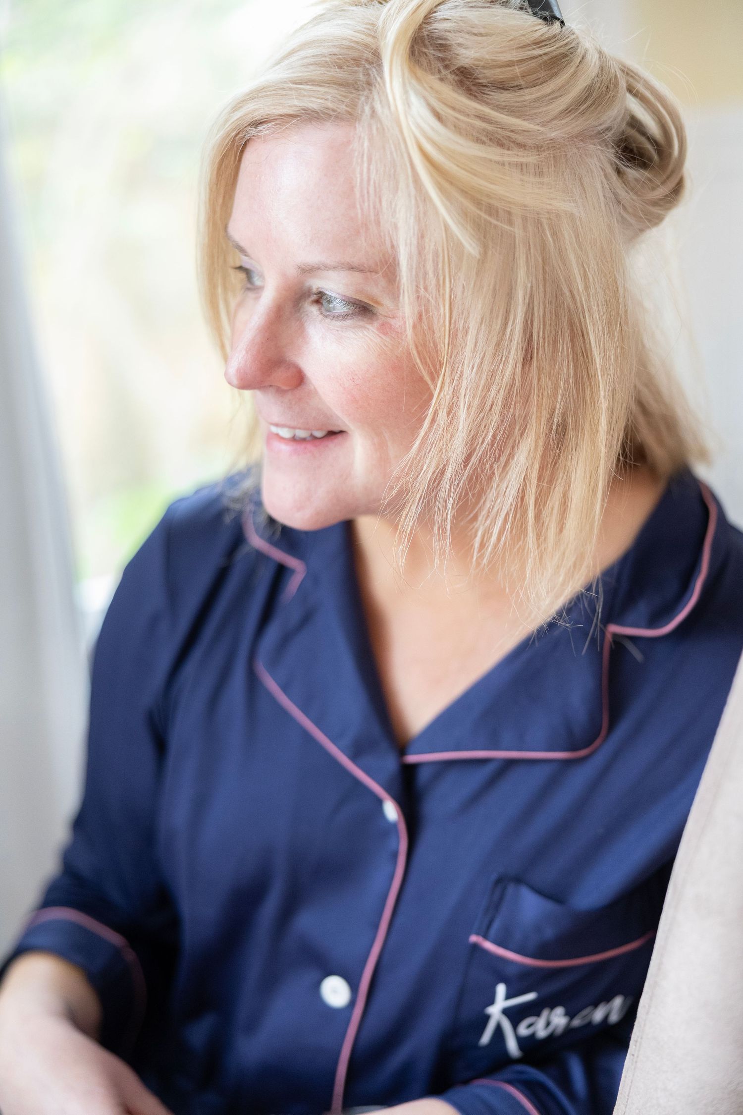 Smiling blonde woman wearing navy blue satin personalized pajamas with pink trim, looking to the side in soft natural light.