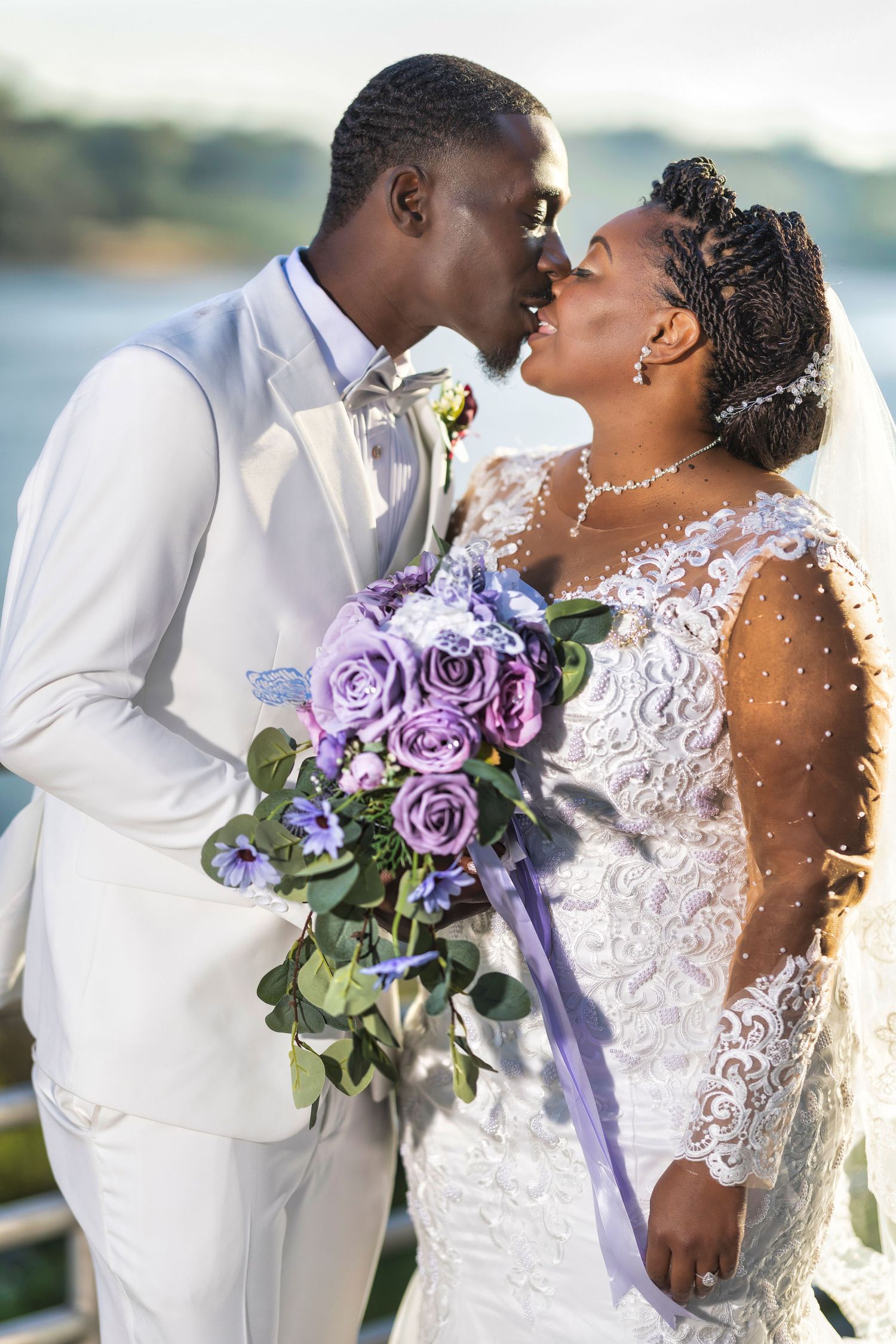 A bride and groom share a romantic kiss outdoors, she holds a purple rose bouquet, wearing lace gown, he in white suit.