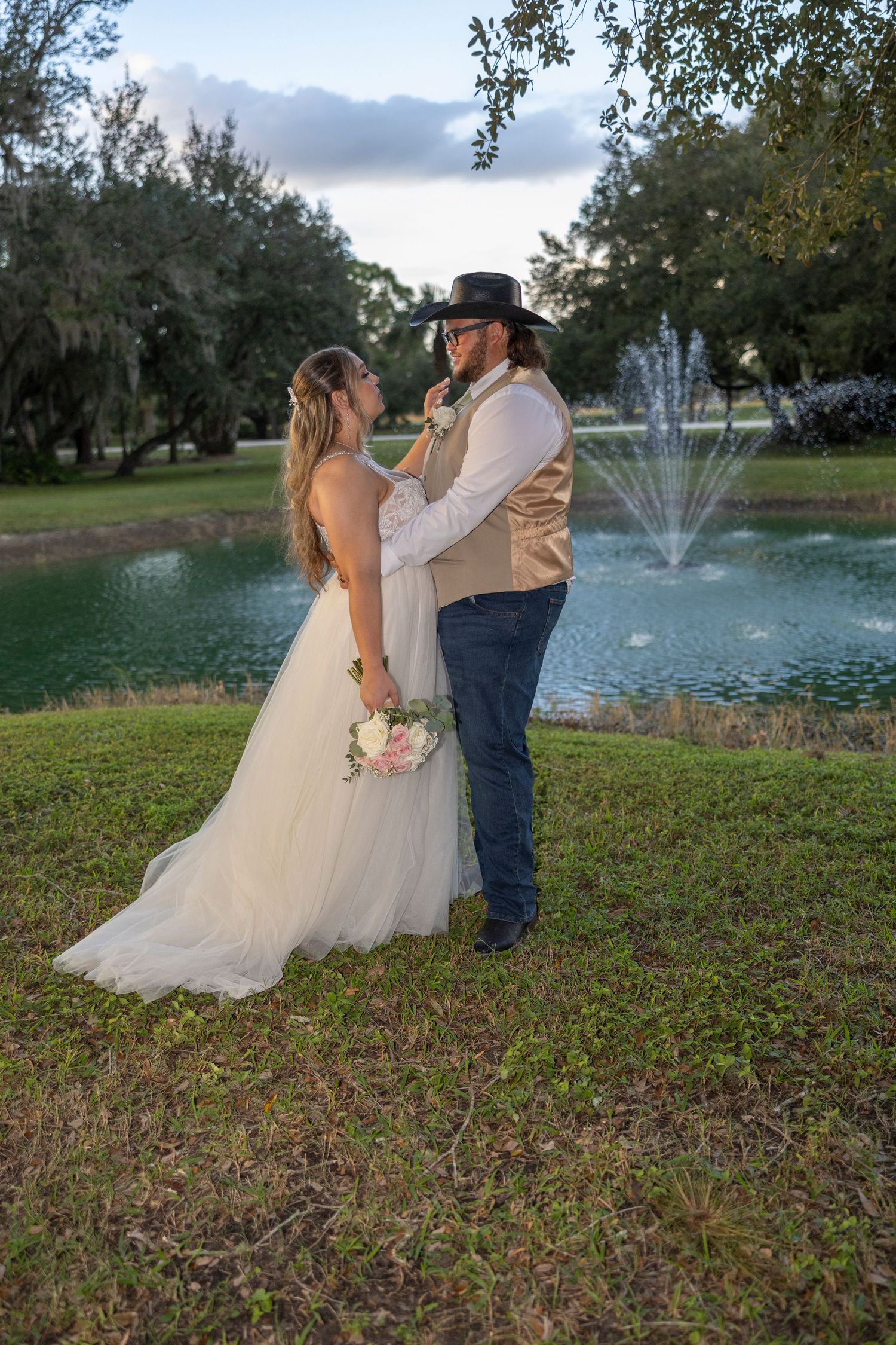 A couple in wedding attire stands by a fountain in a scenic outdoor garden setting at sunset.