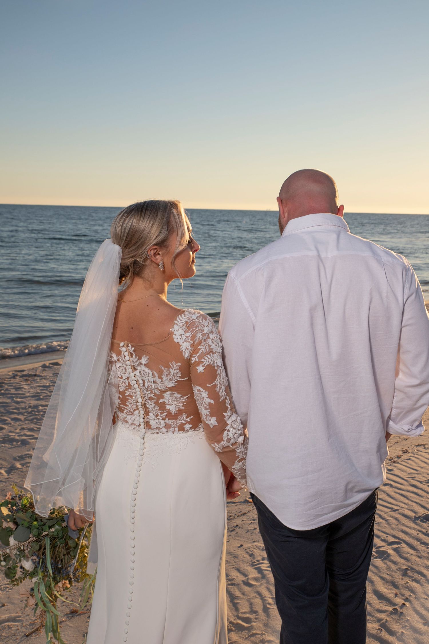 A couple stands together on a beach at sunset, with the bride wearing a white lace wedding dress and veil.