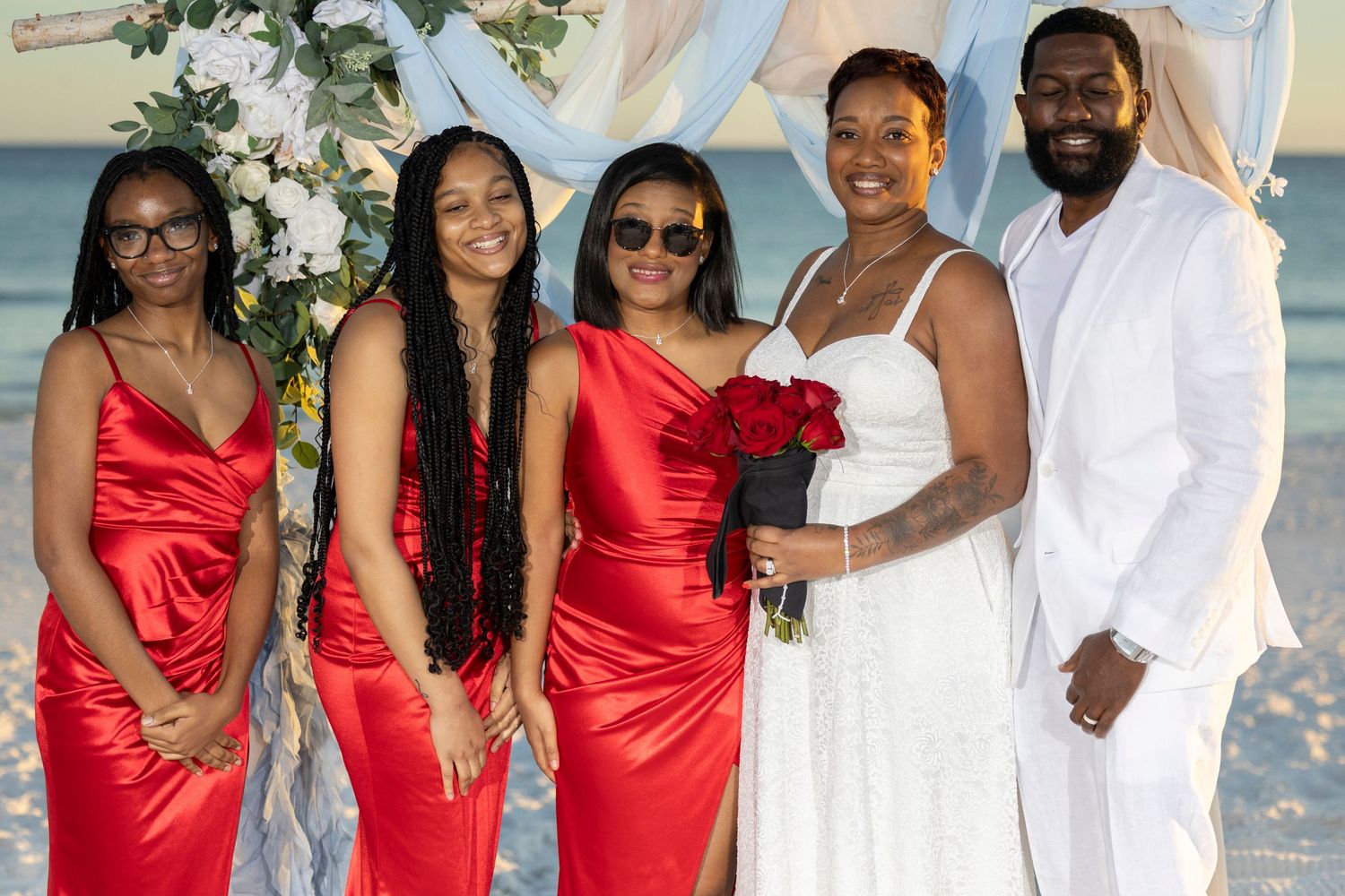 Wedding party in white and red attire poses together on a beach during sunset ceremony.