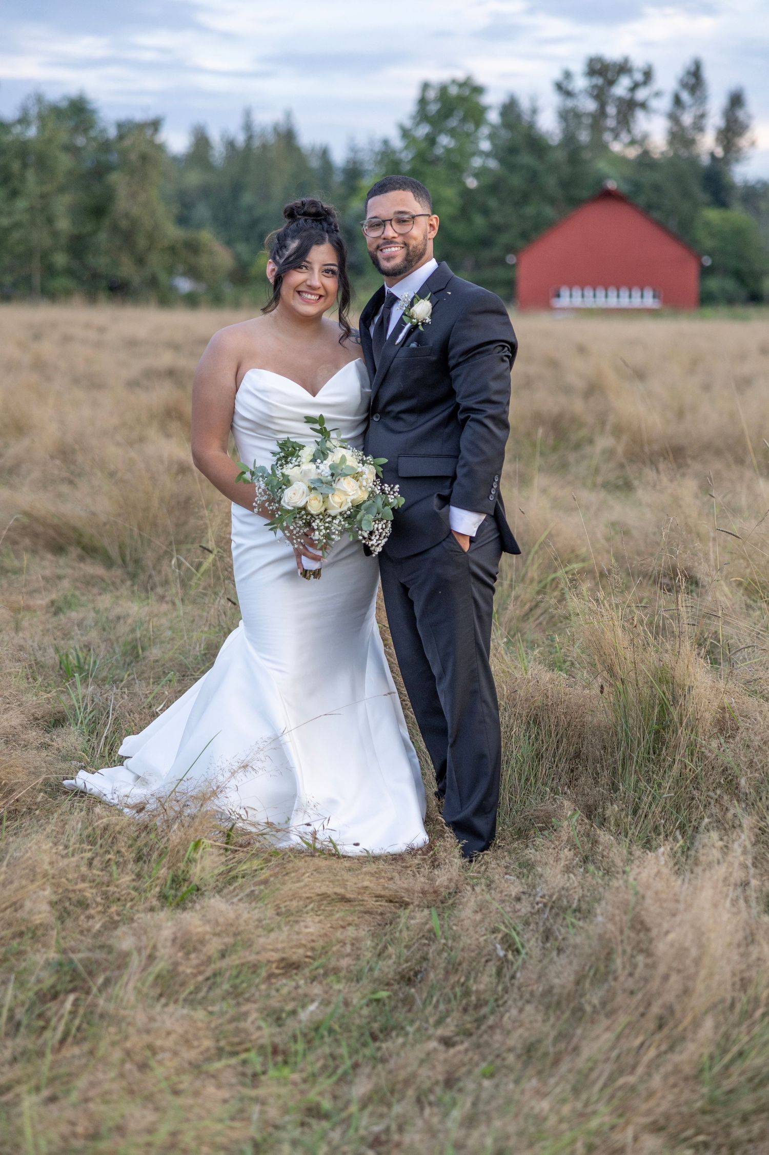 A newlywed couple poses in a rustic field with a red barn in the background on their wedding day.
