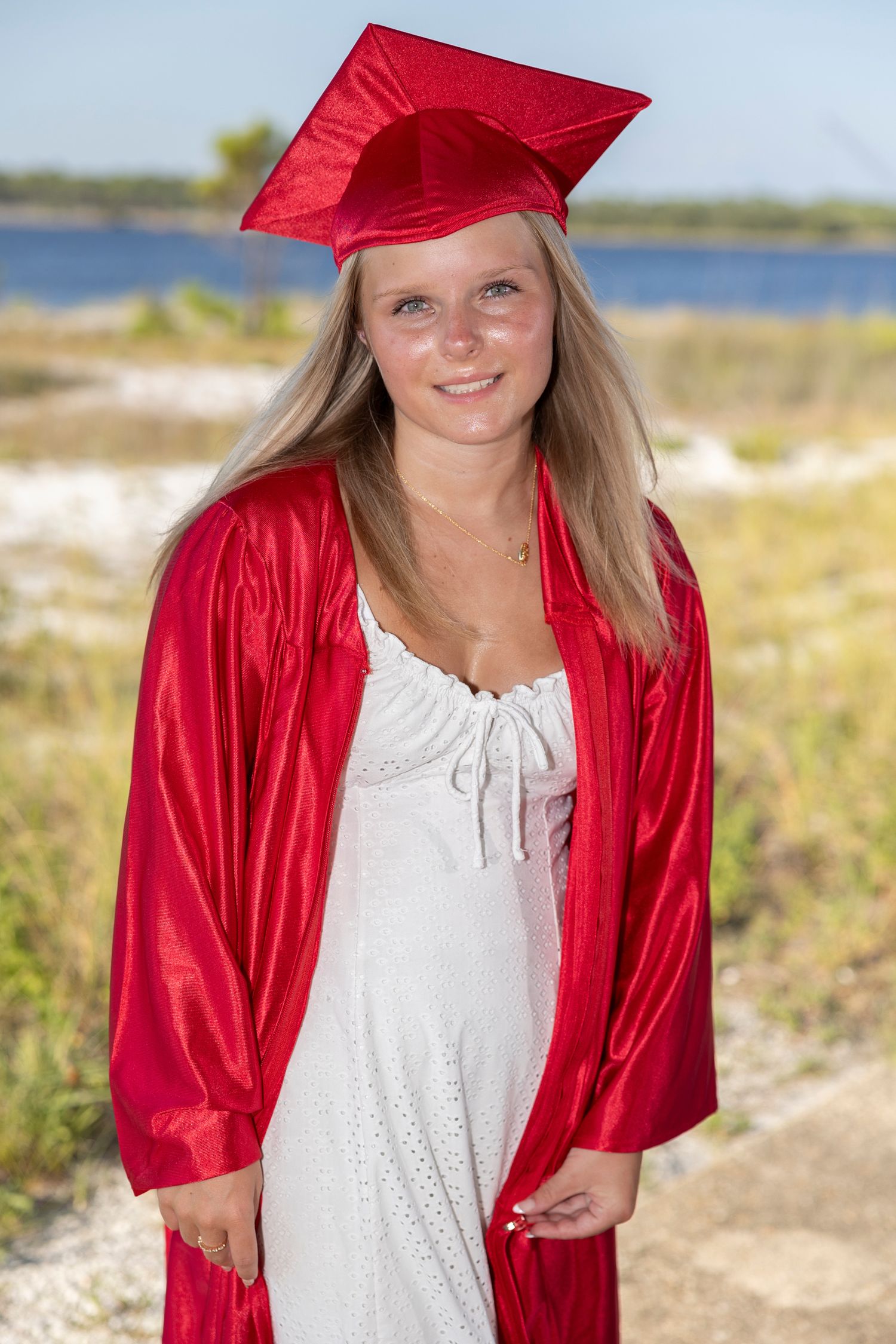 Graduate in bright red cap and gown poses outdoors against coastal landscape on sunny day.