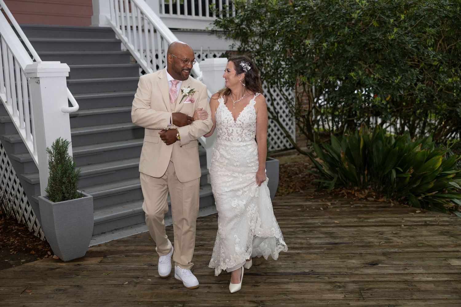 A couple in wedding attire walks together on a wooden deck surrounded by greenery and white railings.