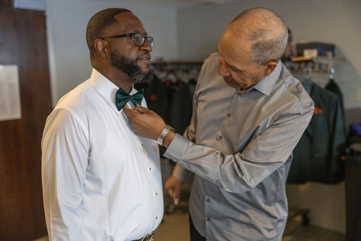 A person helps adjust a green bow tie on someone wearing glasses and a white dress shirt.