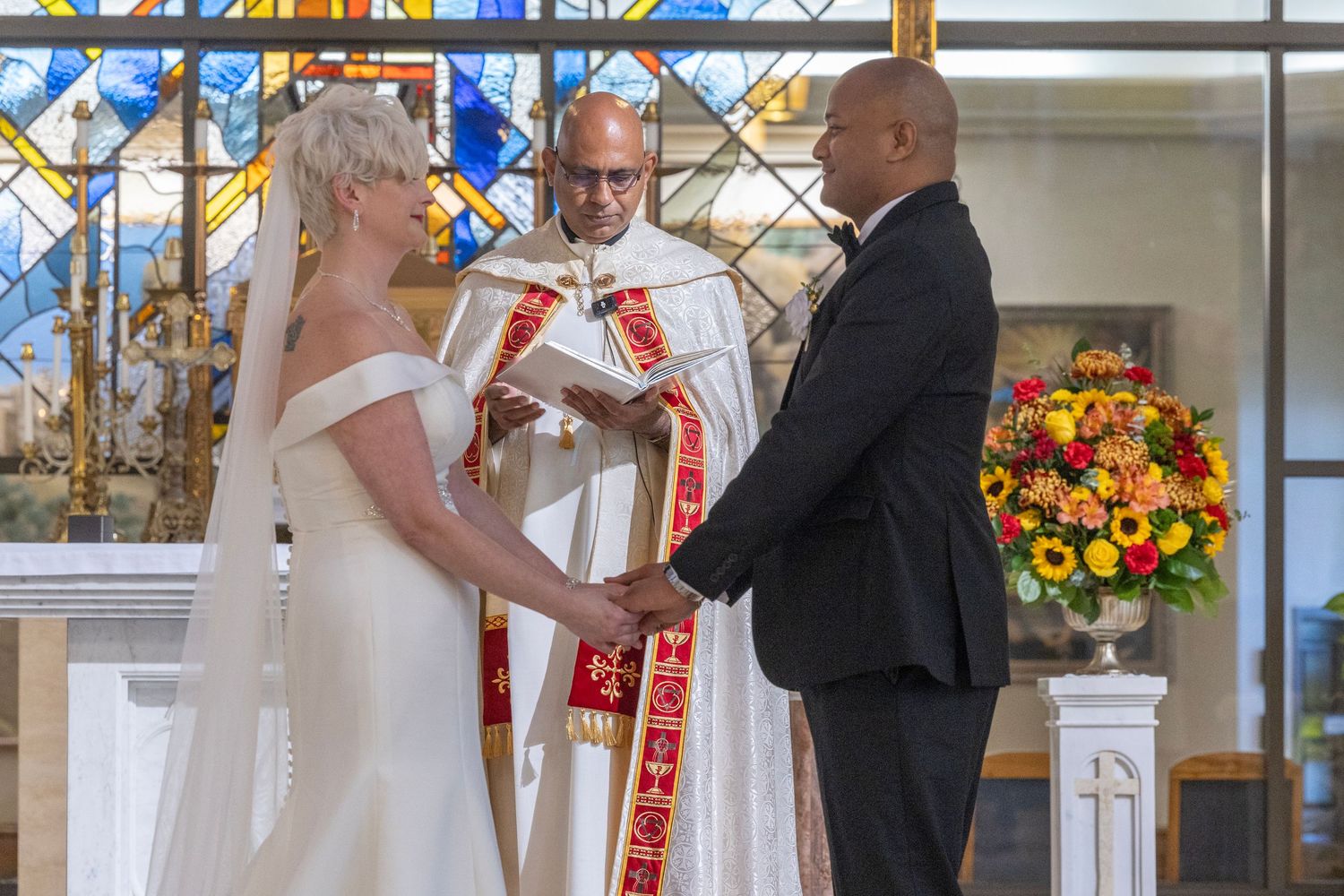 A wedding ceremony takes place in a church with stained glass windows and colorful floral arrangements.